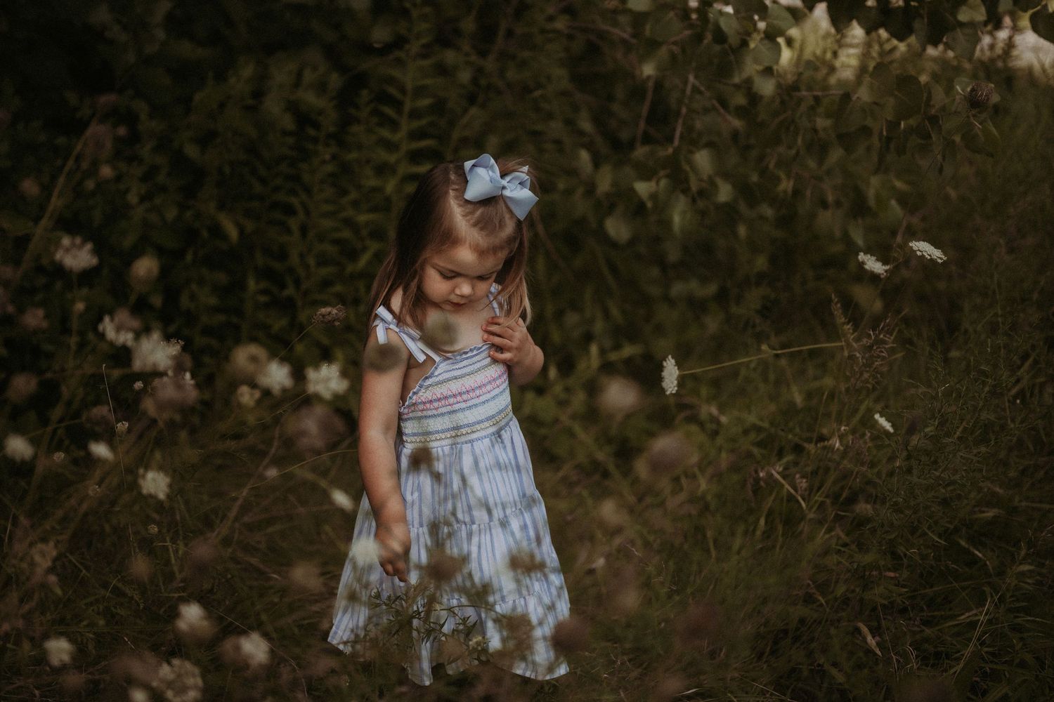 A young child in a light summer dress stands among wildflowers and natural foliage.