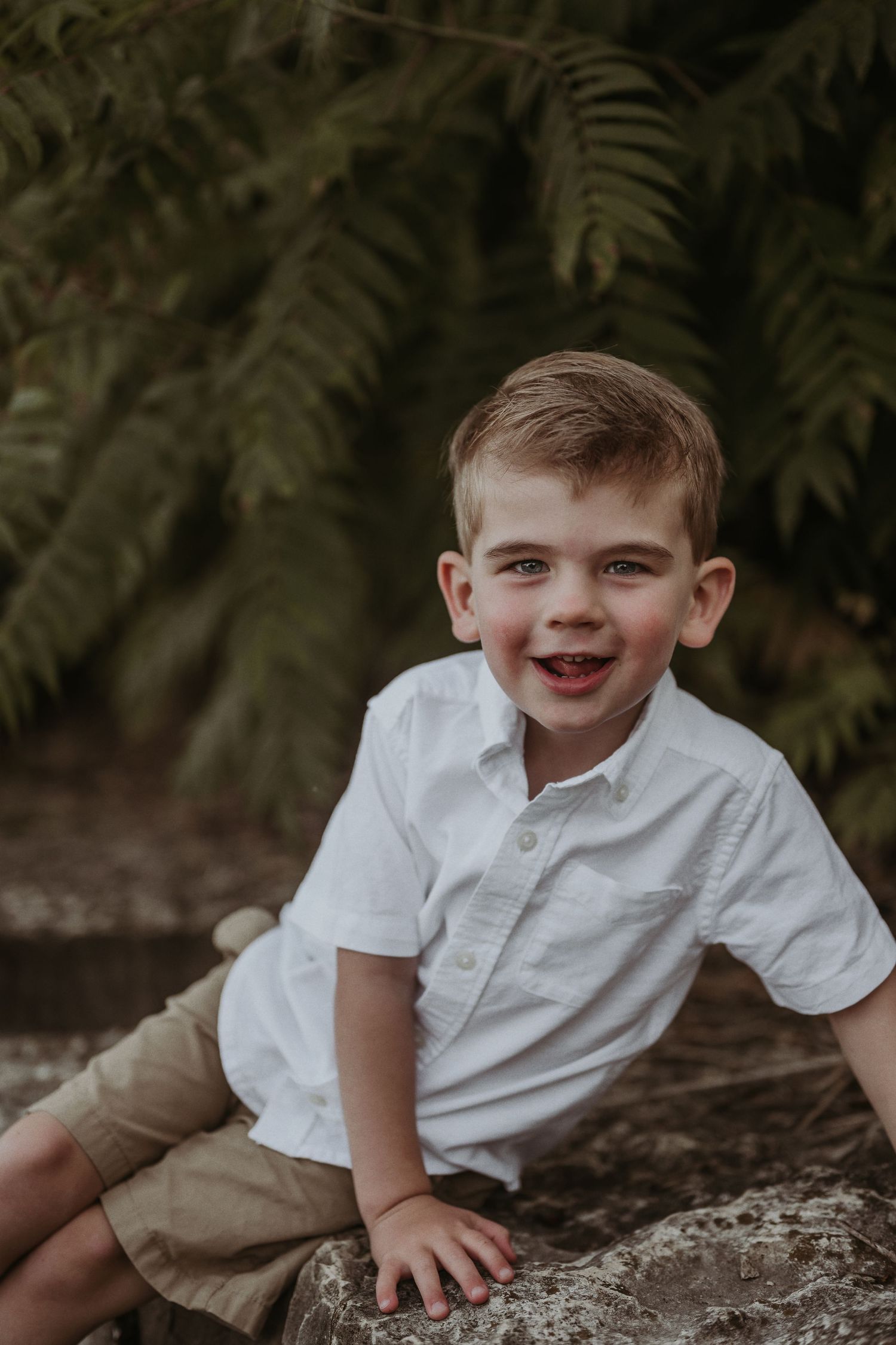 A young child in a white shirt and khaki shorts sits on a rock surrounded by lush foliage.
