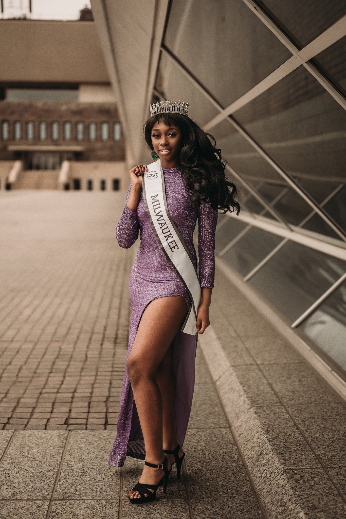 Pageant winner in sparkling purple gown and crown poses near modern architecture.