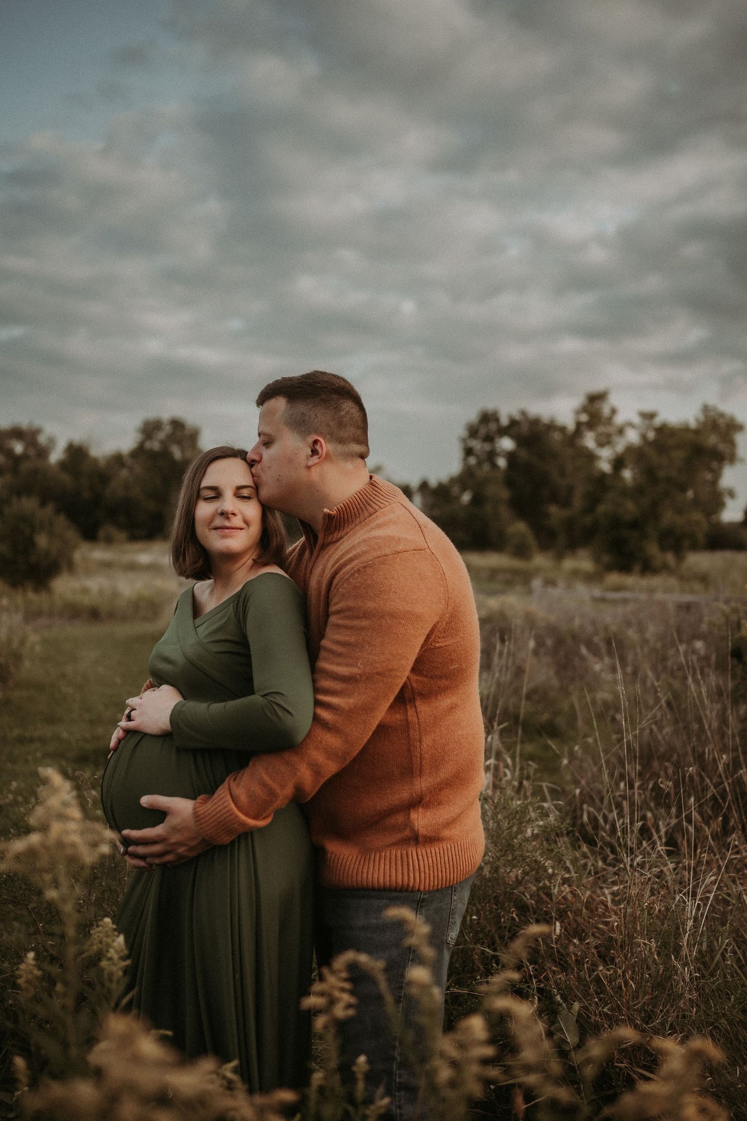 Intimate maternity moment captured in field as couple embraces under cloudy sky.