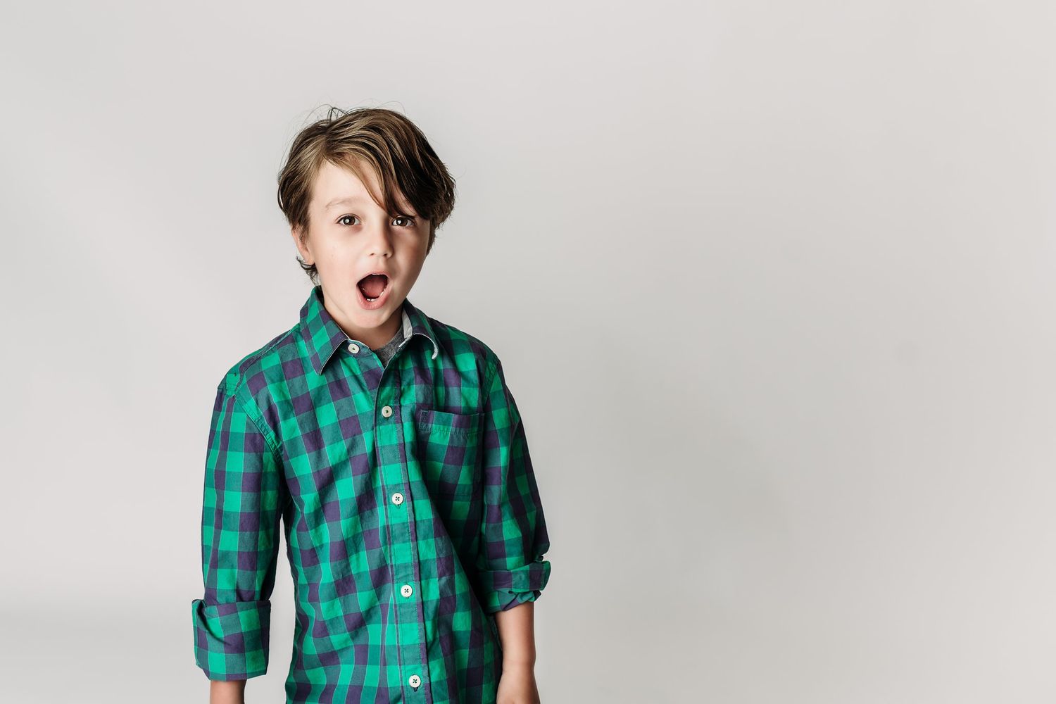 Child in green plaid shirt making surprised expression on white background.