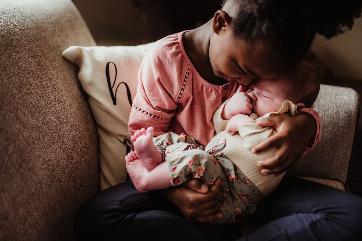 Person in pink sweater gently holding and looking down at a sleeping baby on couch.