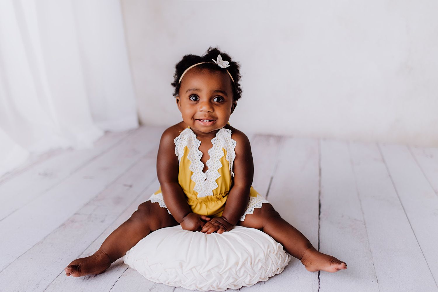 Happy baby in yellow outfit sitting on white cushion during photo shoot.