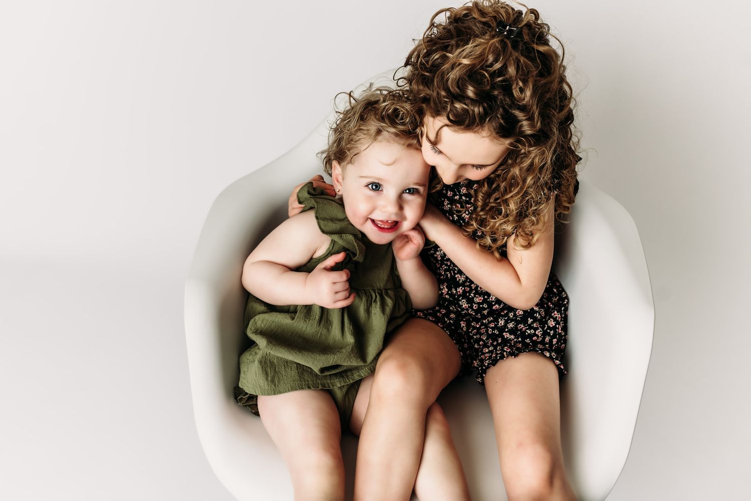 Two children share laughter while cuddled together in a white modern chair.