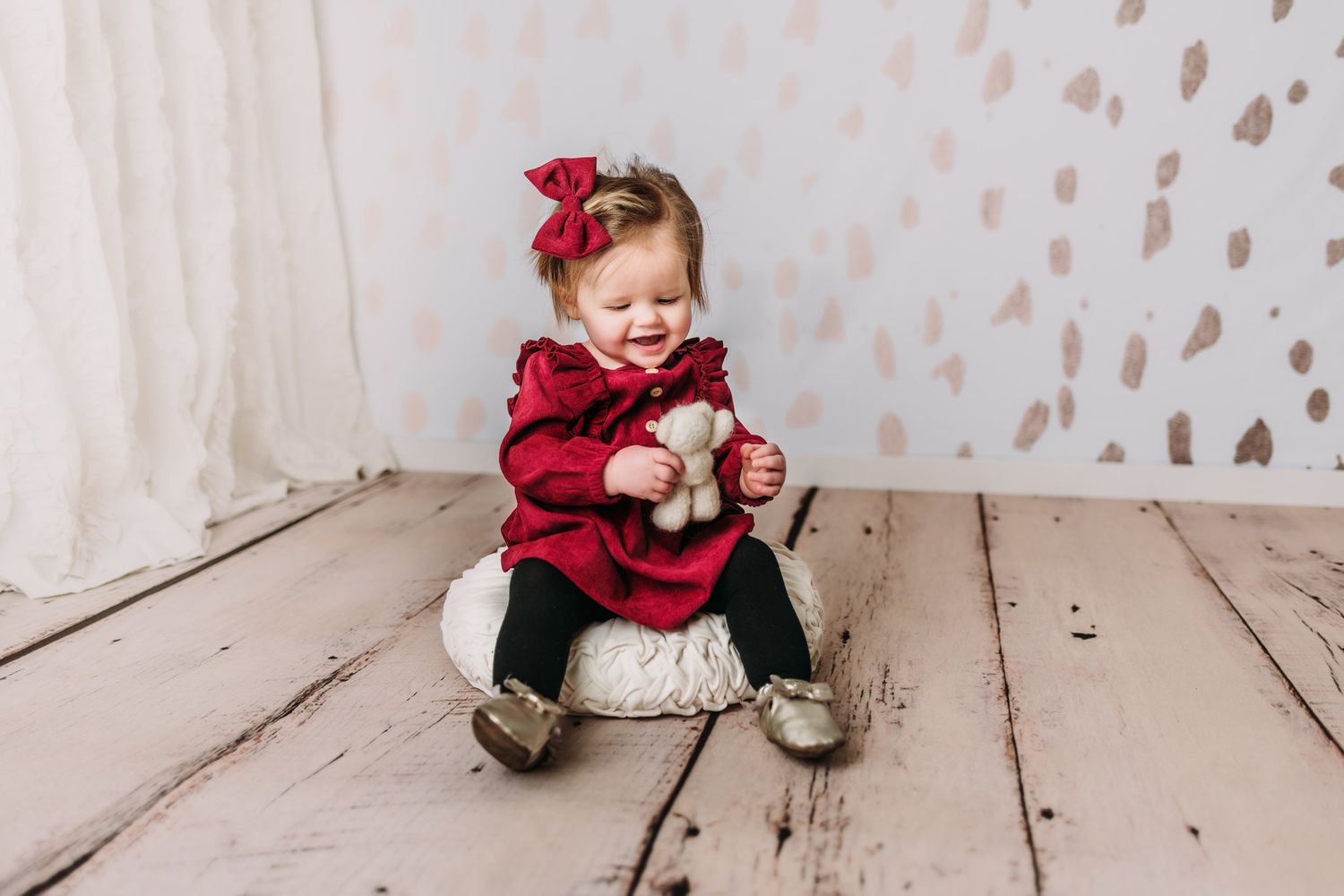 Baby in red dress holding teddy bear on white wooden floor.