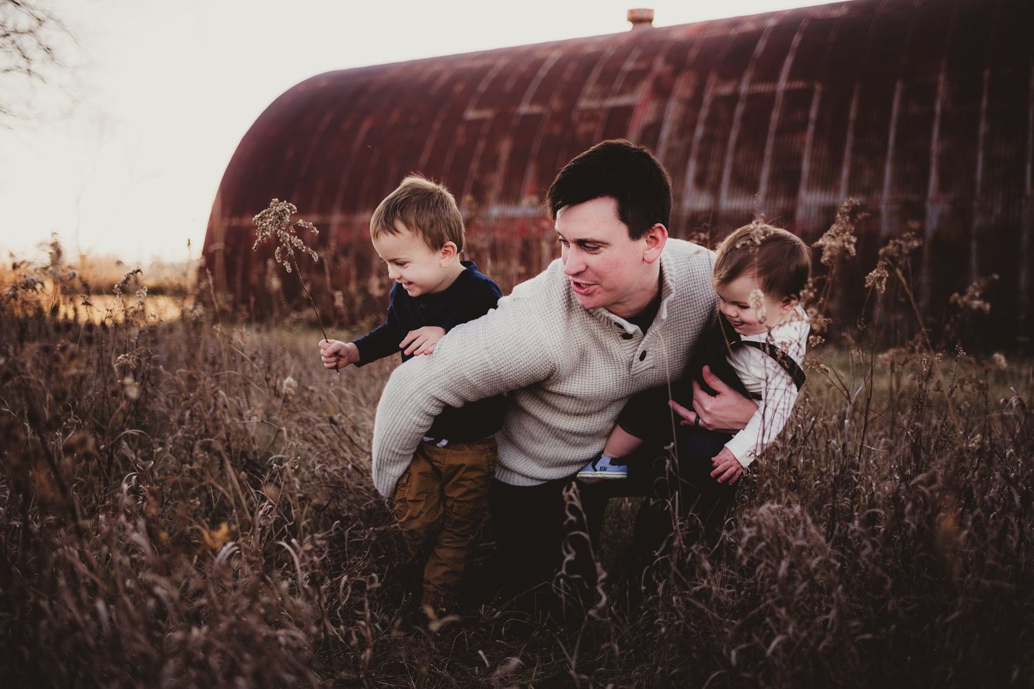 Playful family interaction in tall grass with rustic barn backdrop.