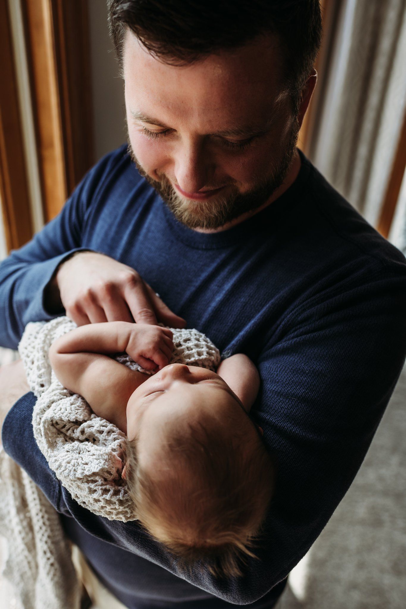 Parent in navy sweater tenderly holding a sleeping infant wrapped in a knit blanket by a window.