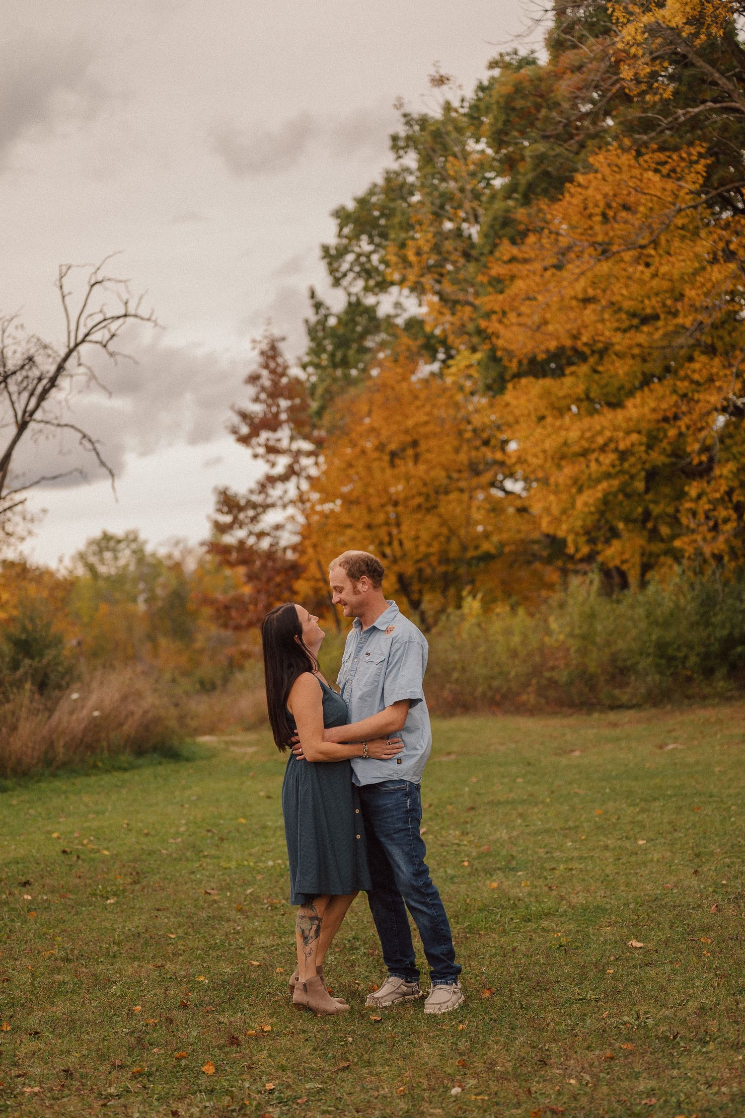 A couple shares a romantic moment in a scenic autumn field with orange and yellow foliage in the background.