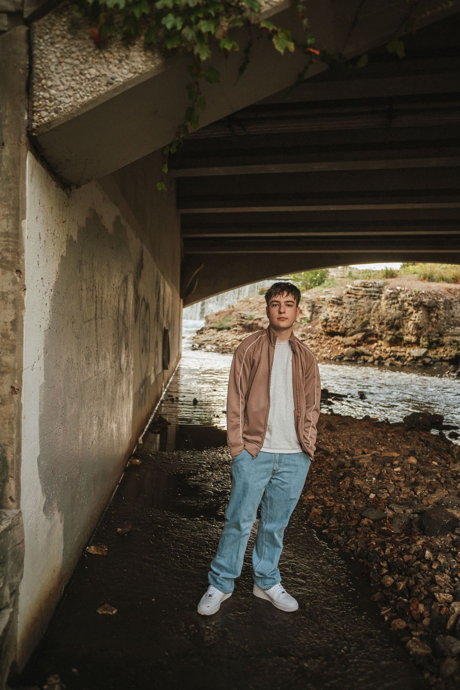 A figure stands beneath a concrete bridge structure near fallen leaves and a flowing stream.