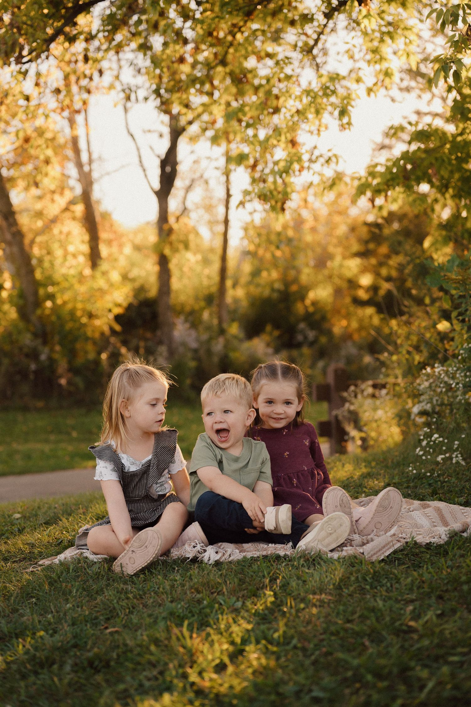 Three children laying on a blanket outdoors during sunset with fall foliage in the background.