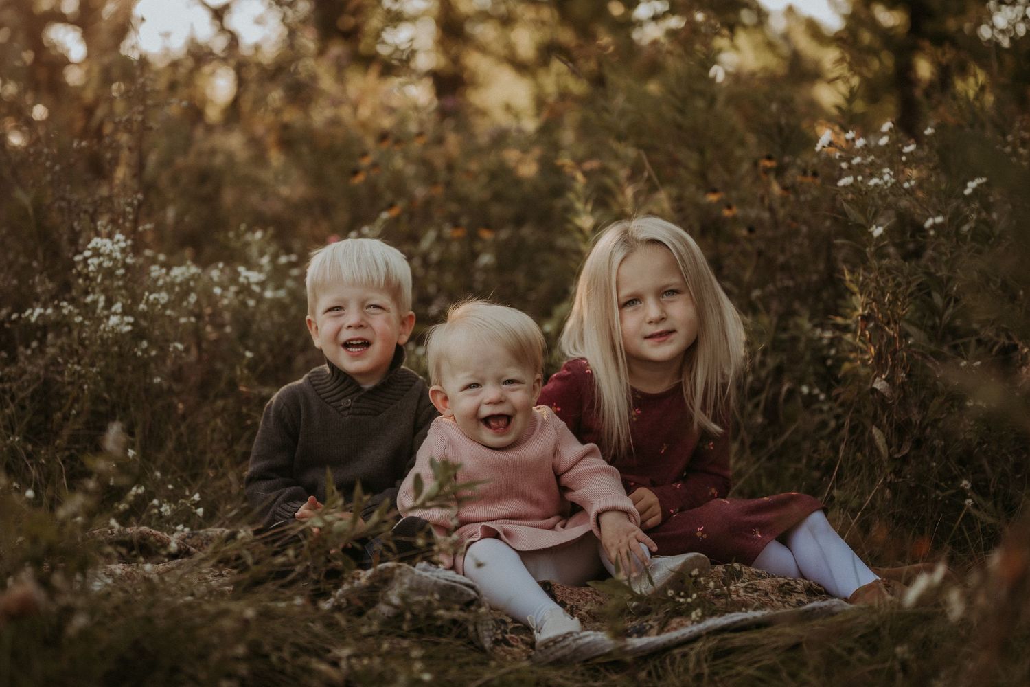 Three children sitting together among wild grass during golden hour photoshoot.