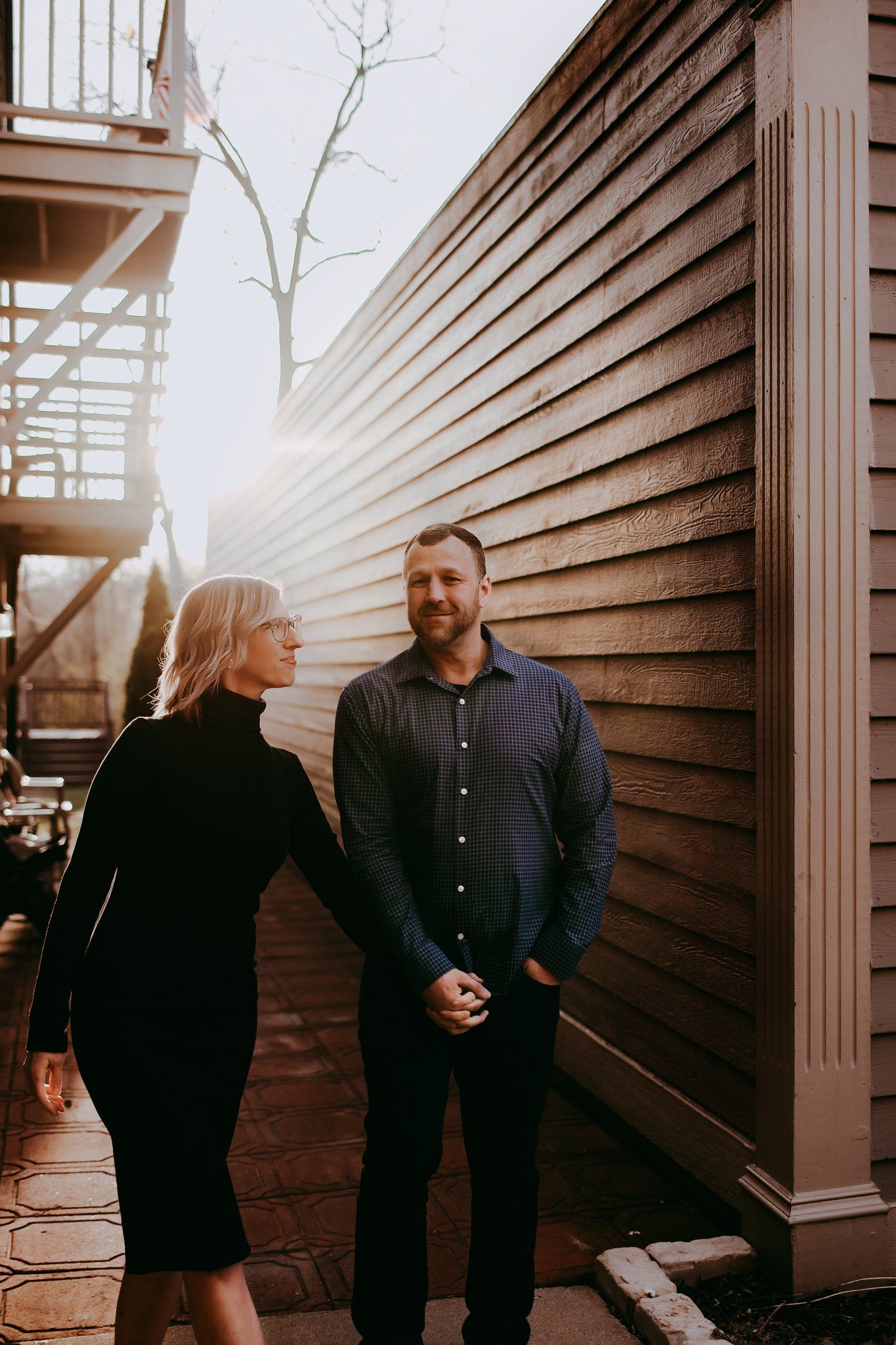 Stylish couple in dark clothing standing between wooden walls during golden hour.