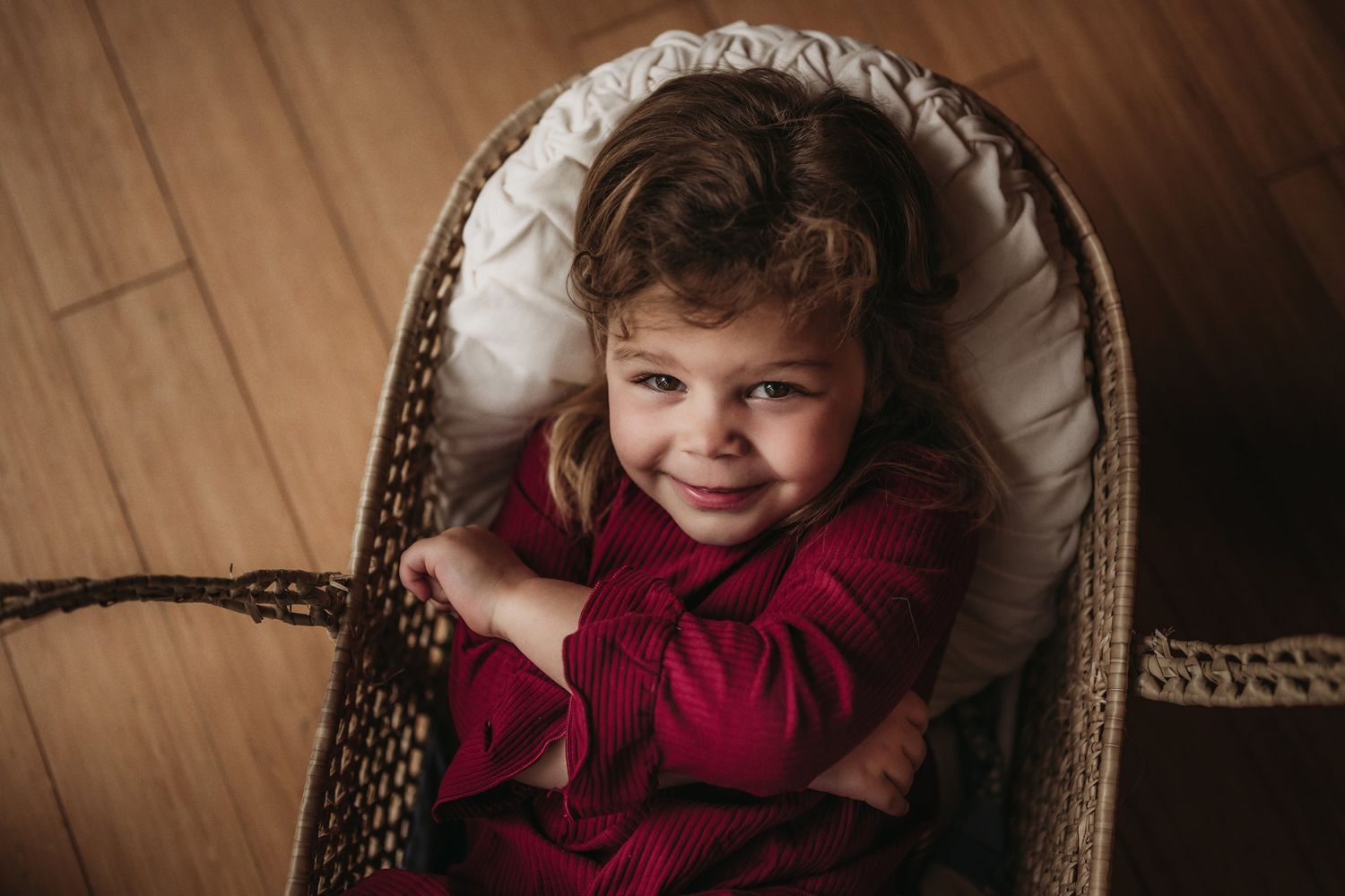 Child in red dress smiles while sitting in woven basket on hardwood floor.