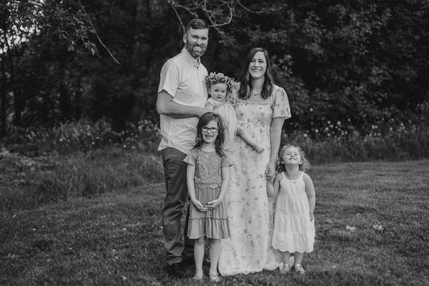 Black and white portrait of family standing together in front of wooded backdrop.