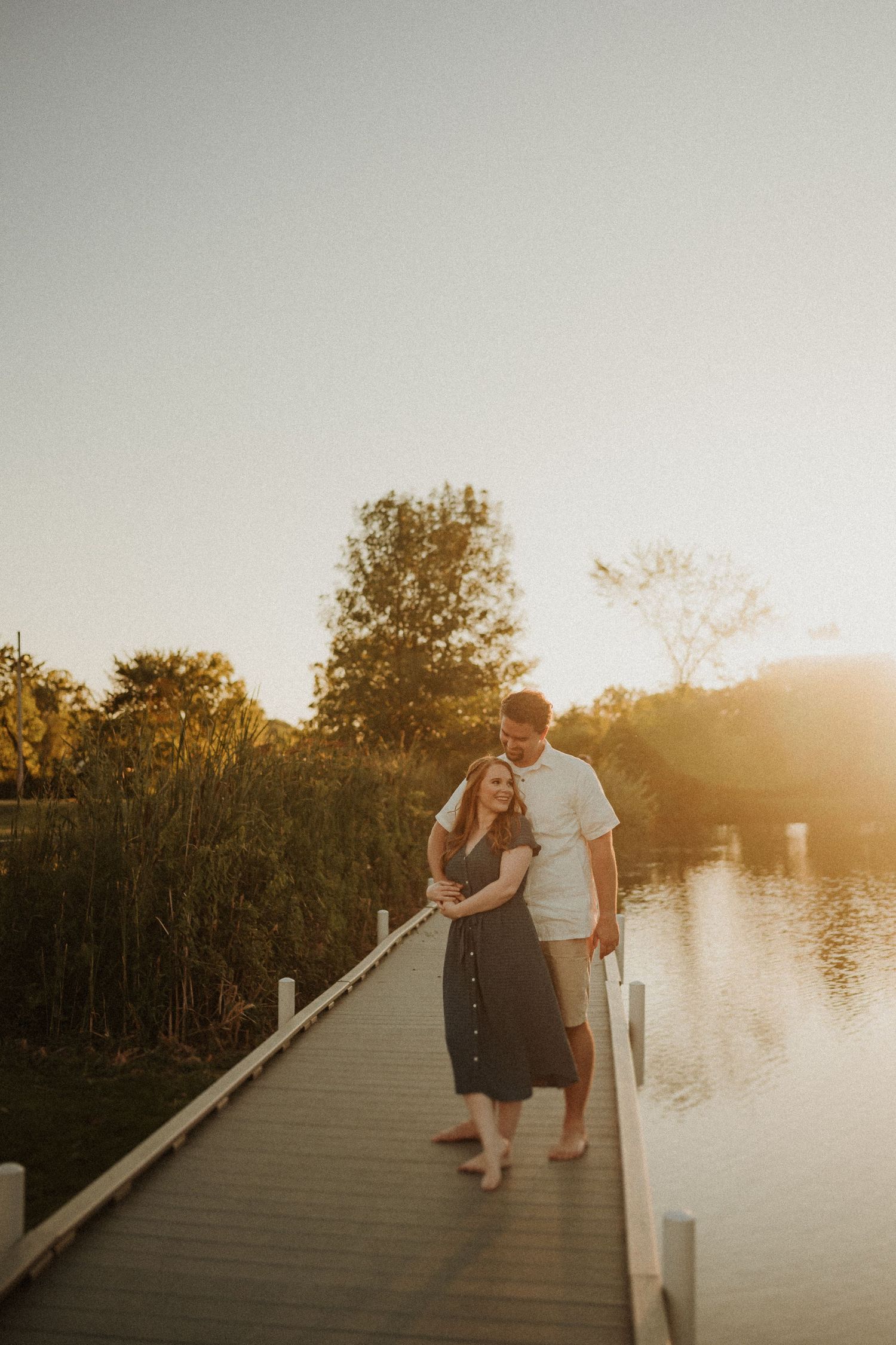 Couple walking on a wooden boardwalk over a lake at sunset with trees in the background.