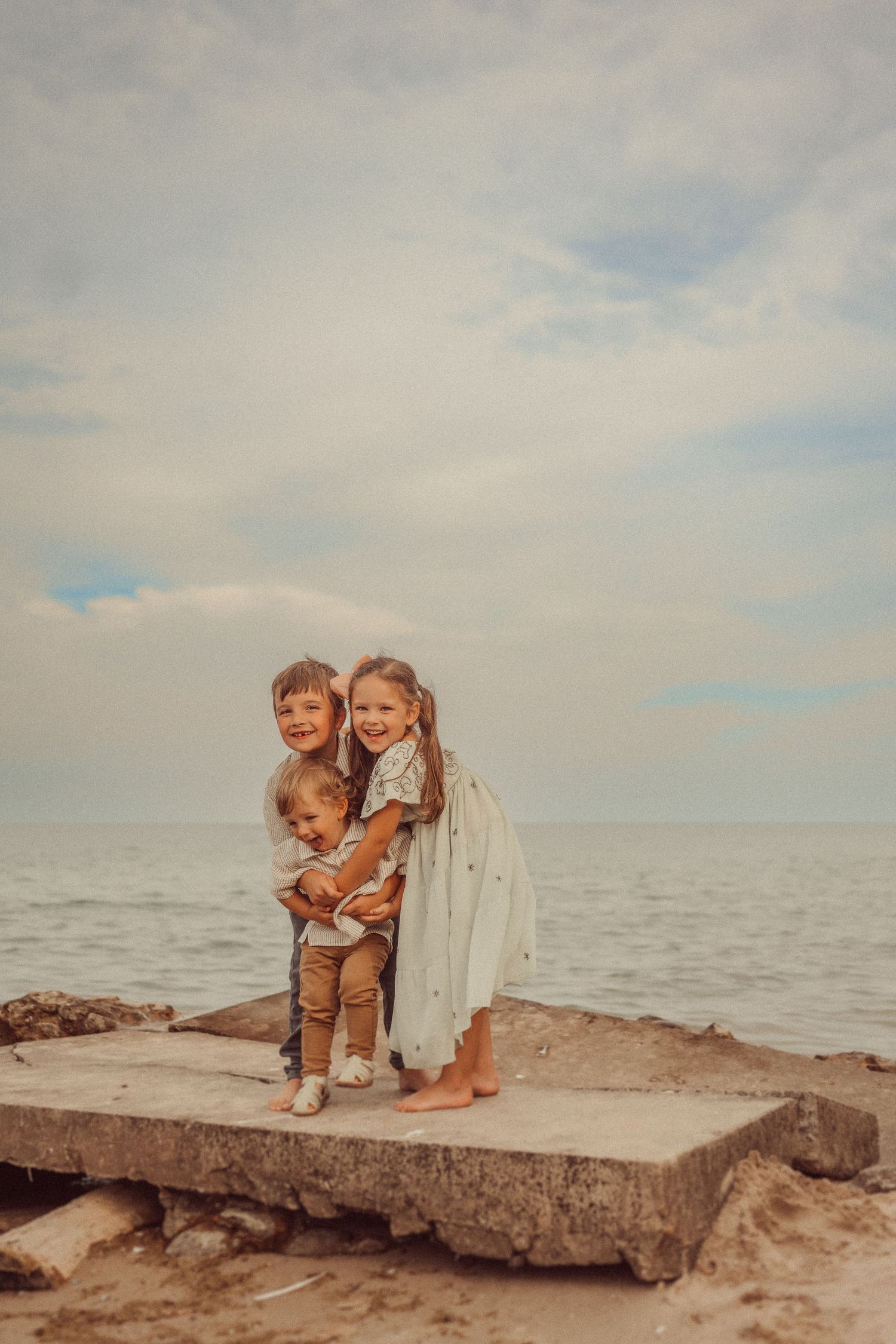 Three children hug while standing on a concrete block at the beach.