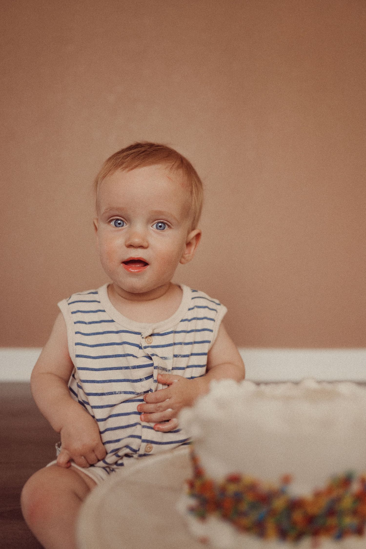 Toddler in striped outfit sitting beside decorated white cake during photo session.