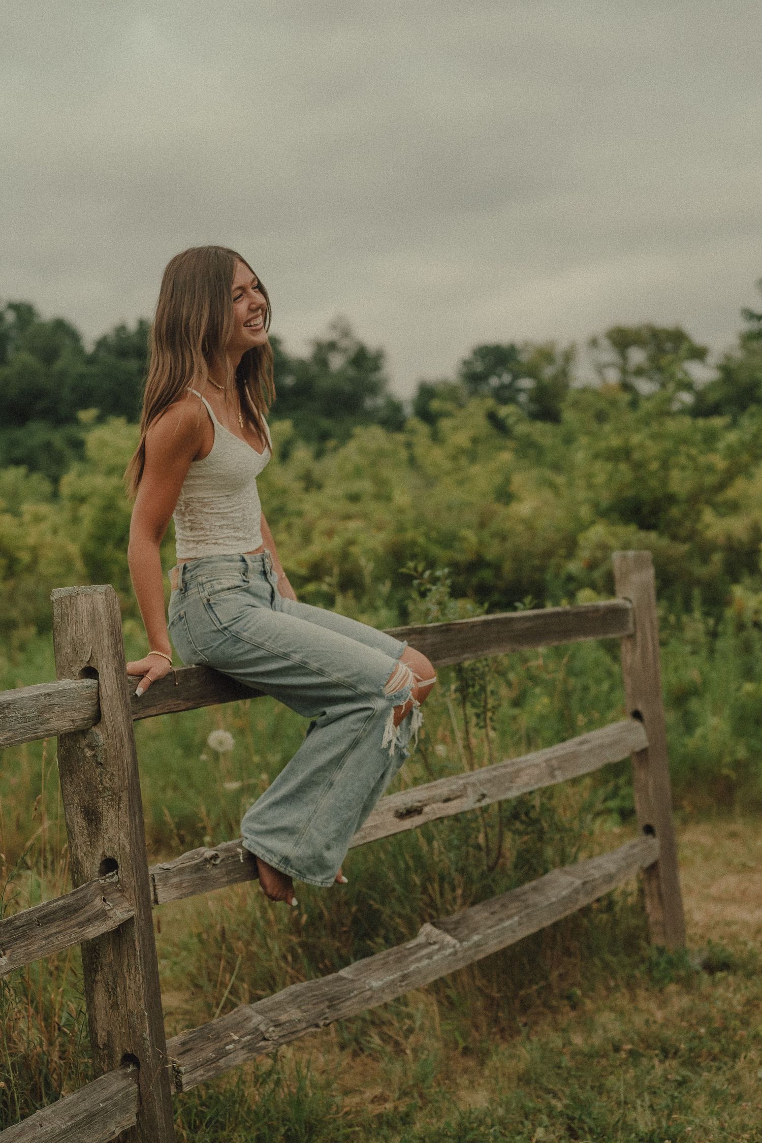 Laughing young woman in white tank and wide-leg jeans perched on a wooden fence in a meadow.