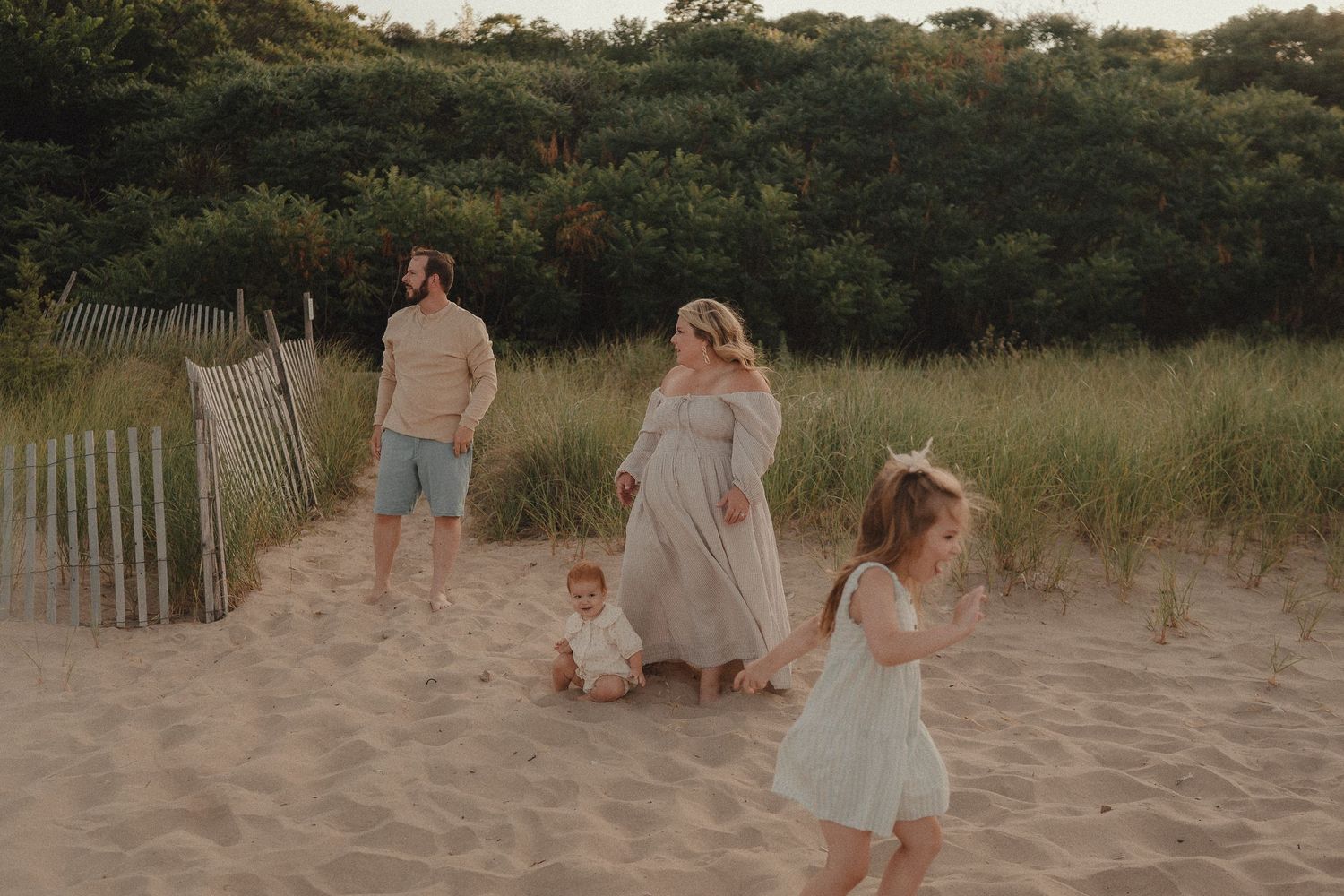 Family of four on a sandy dune, with parents watching their toddler sit and young girl in a white dress playing nearby.