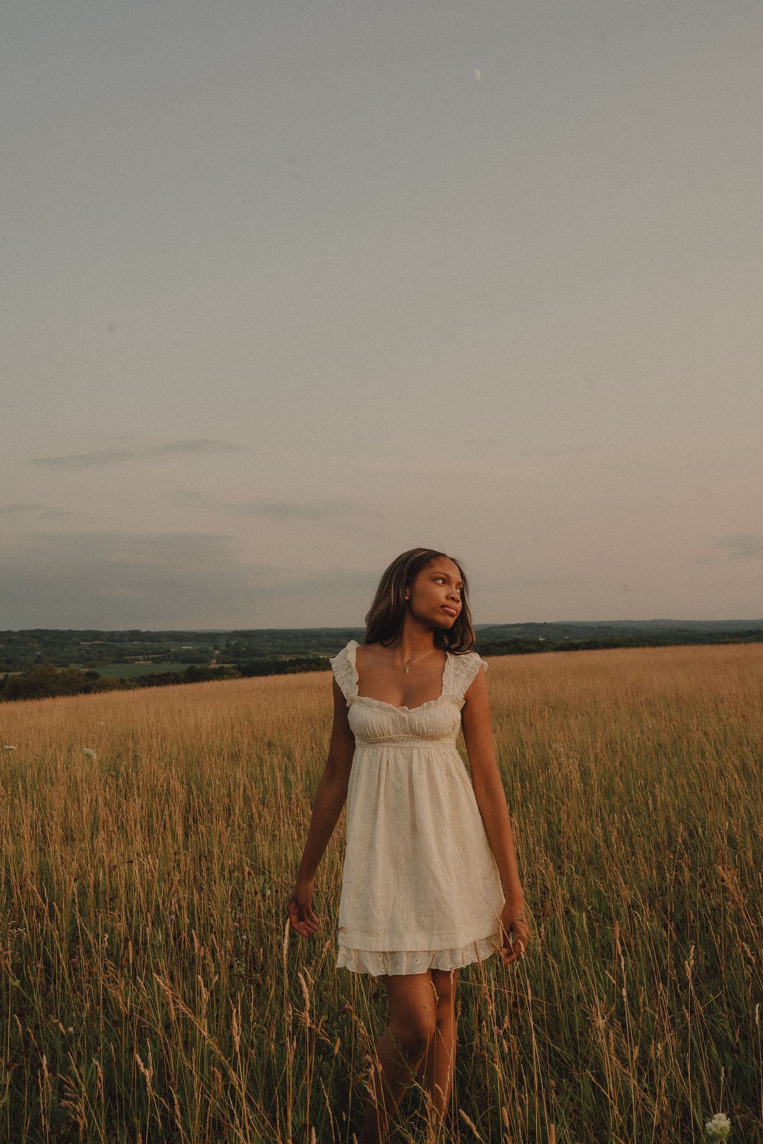 Young woman in white dress stands in a golden field at sunset under a soft evening sky.