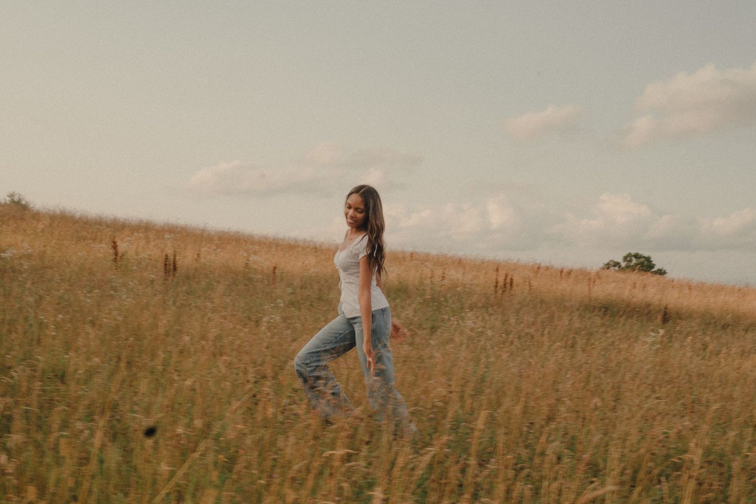 A young woman in a white top and jeans walks through a golden field of tall grass under a cloudy sky.