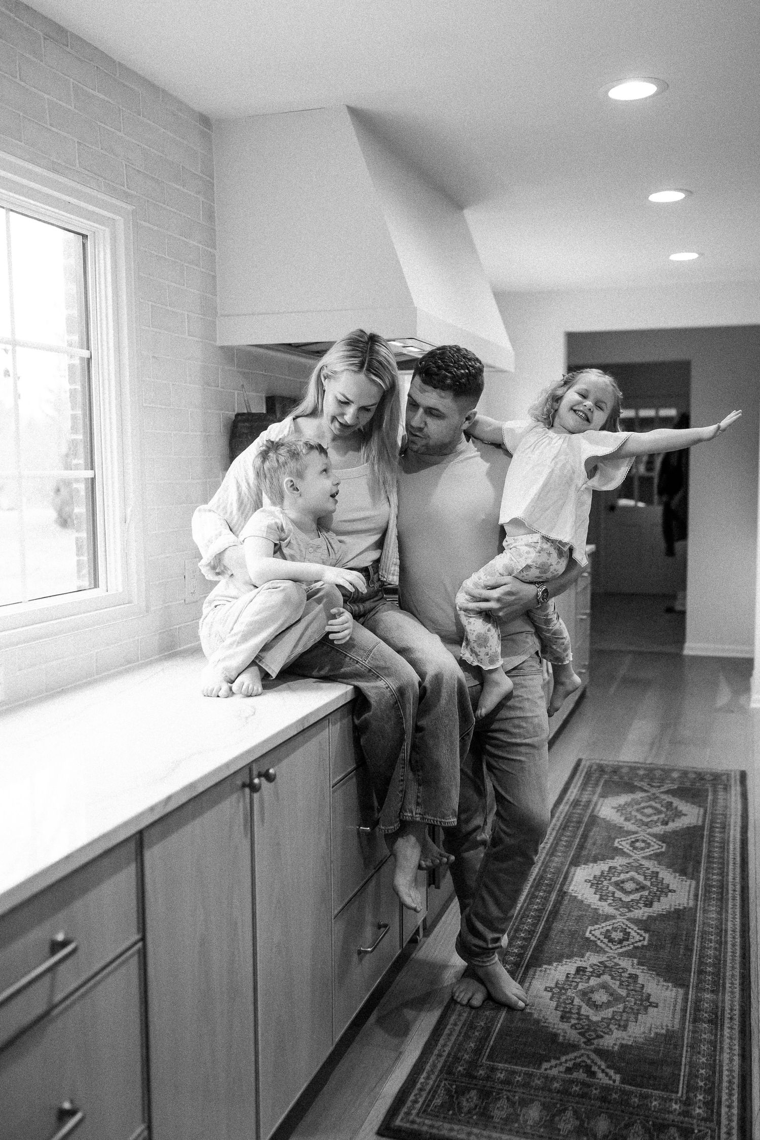 Black and white family portrait of parents with three children sharing tender moment on kitchen counter.