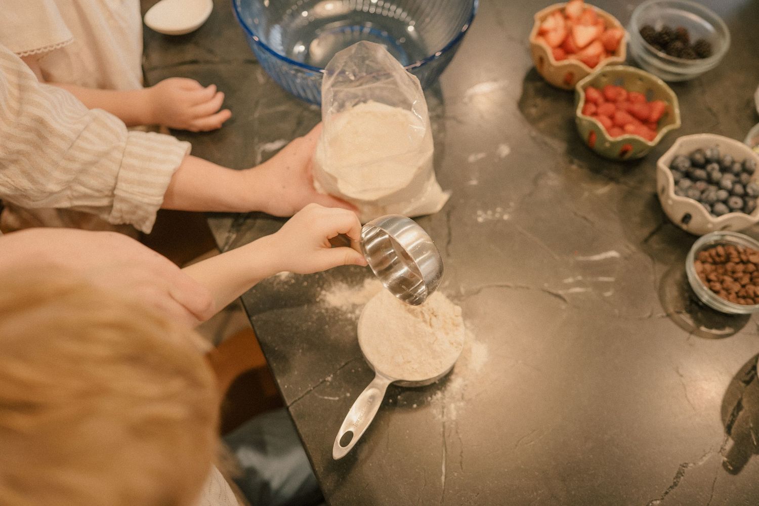 Children baking with flour on a dark countertop, with bowls of fresh strawberries and blueberries nearby.