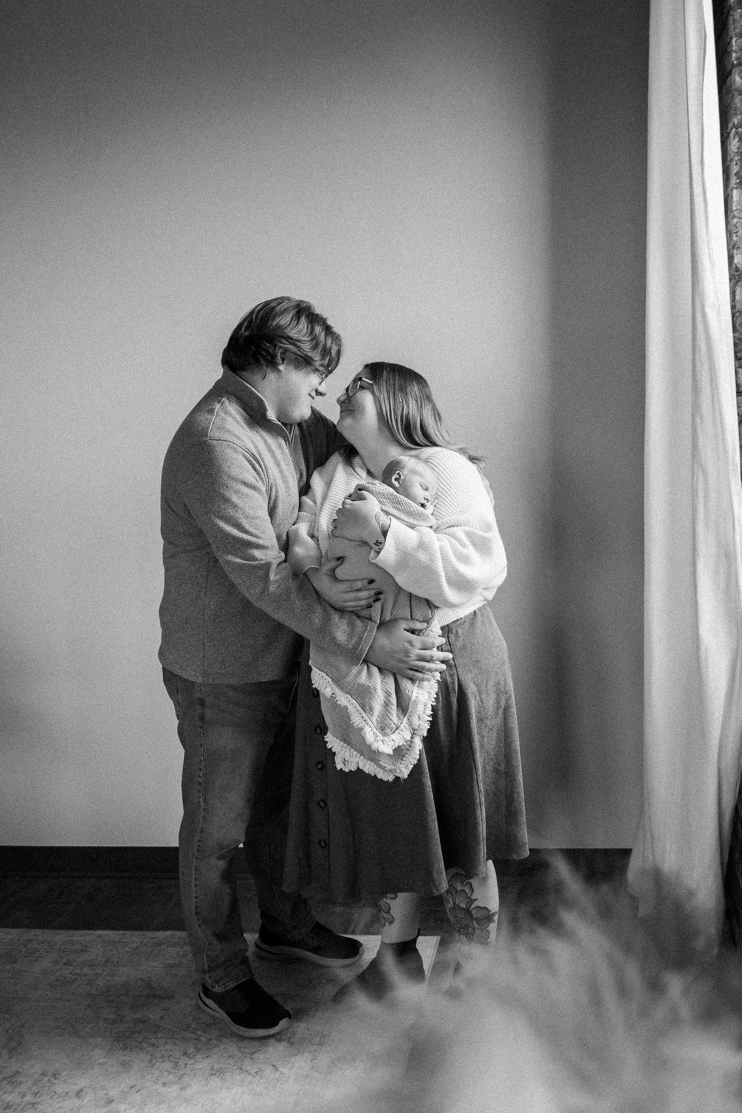 Black and white photo of a couple kissing over their newborn baby wrapped in a white blanket by a window.