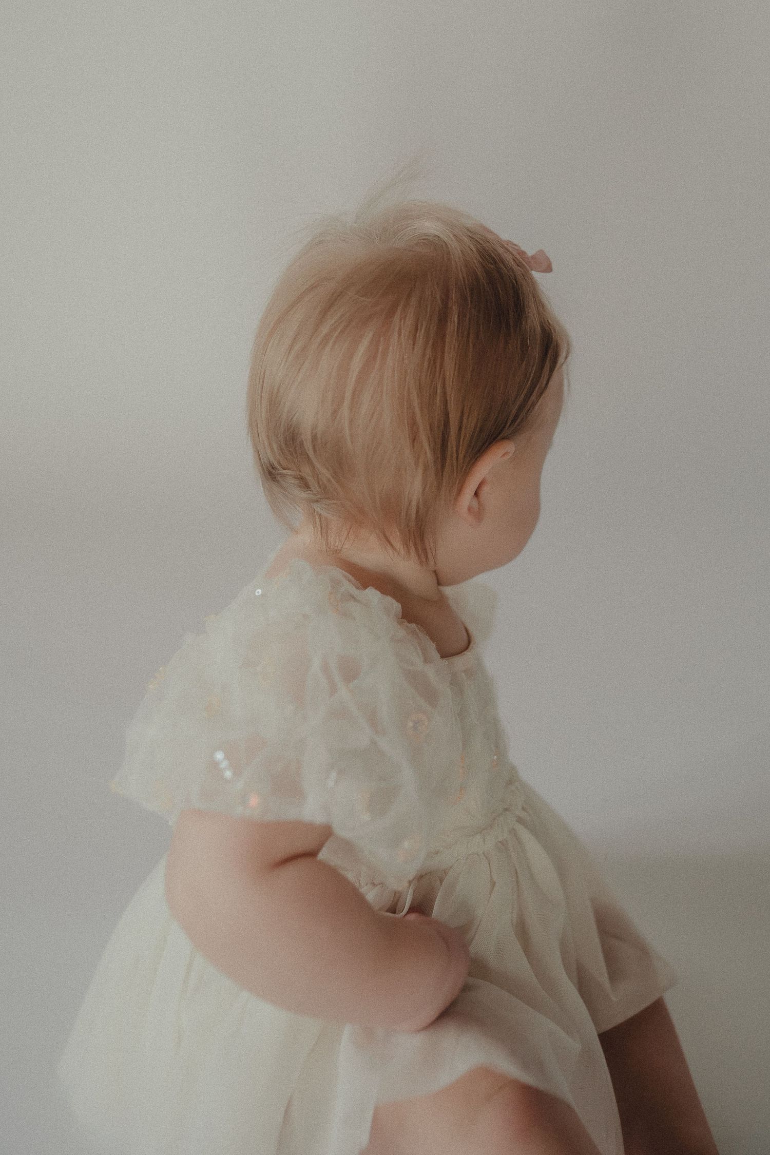 Back view of a baby girl in a white floral tulle dress with a small bow in her fine blonde hair.