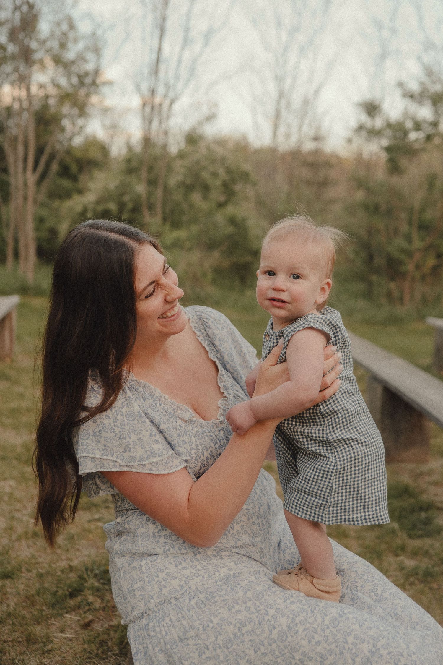 Joyful mother in grey dress lifts her smiling baby girl while seated on a bench in a lush green park.