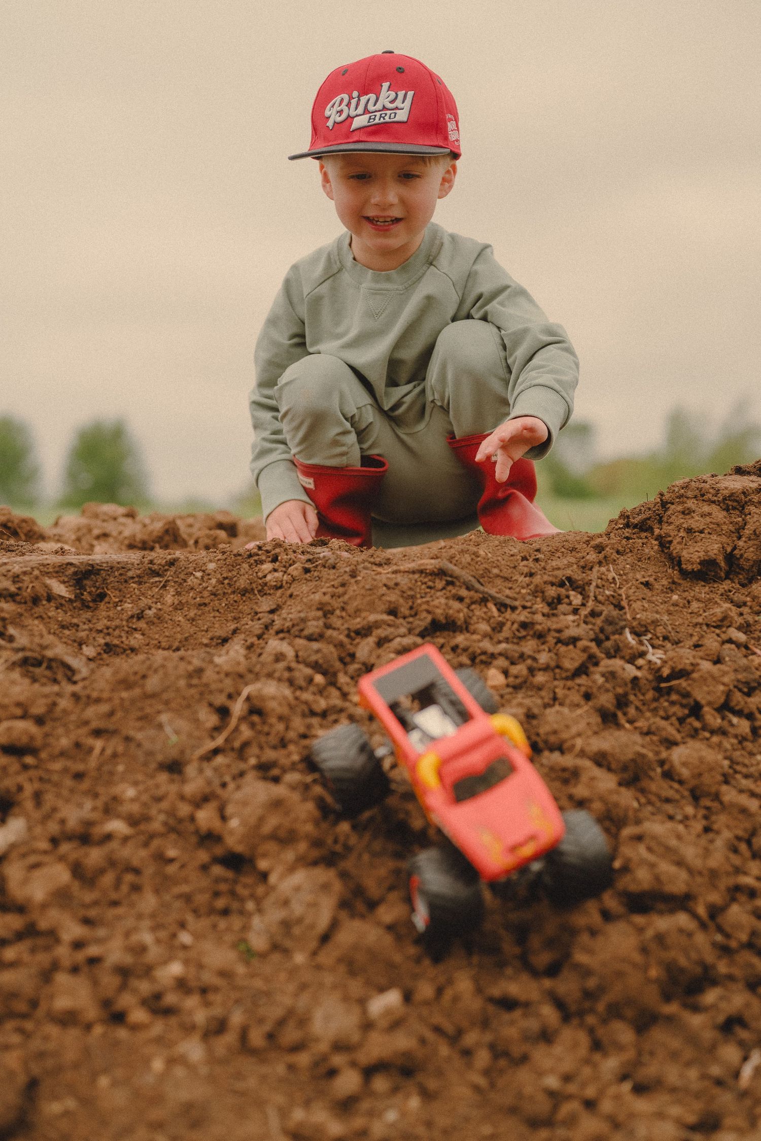 Smiling young boy in red cap crouching in dirt playing with orange monster truck toy outdoors.