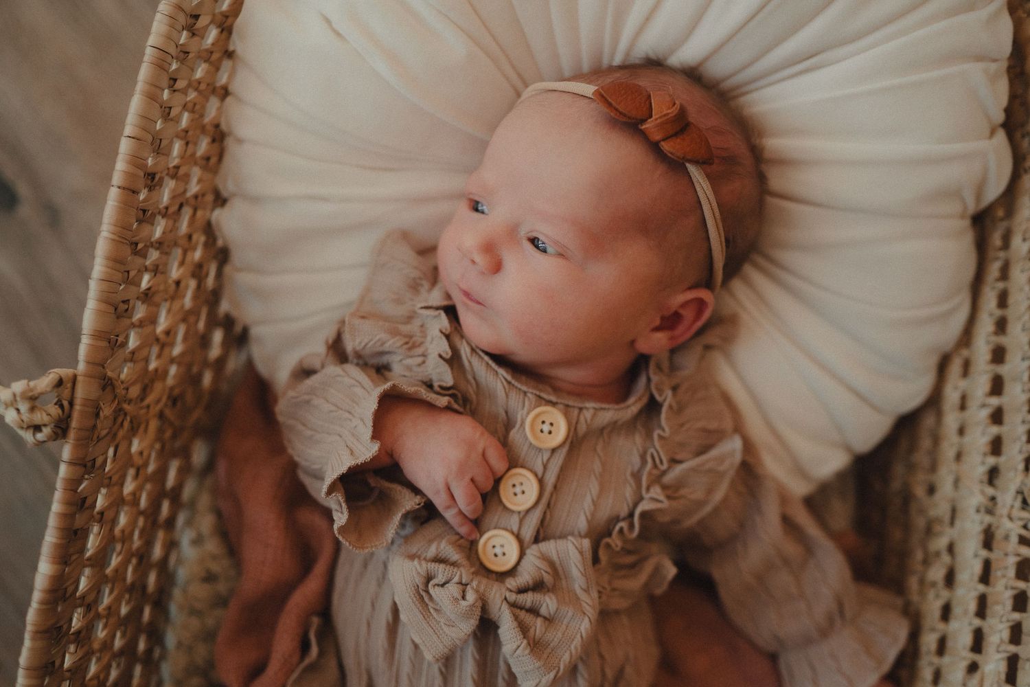 Newborn baby wearing cream colored outfit rests on soft white fabric in vintage wicker basket.