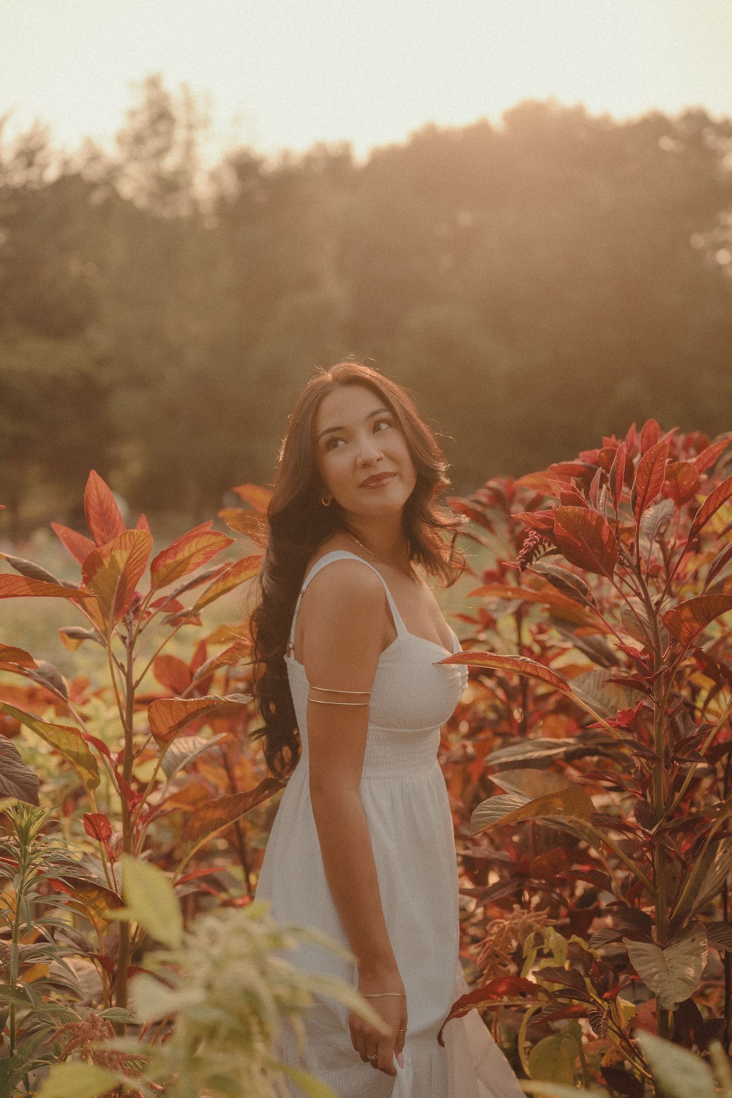 Young woman in white dress among vibrant red foliage bathed in golden sunset light.
