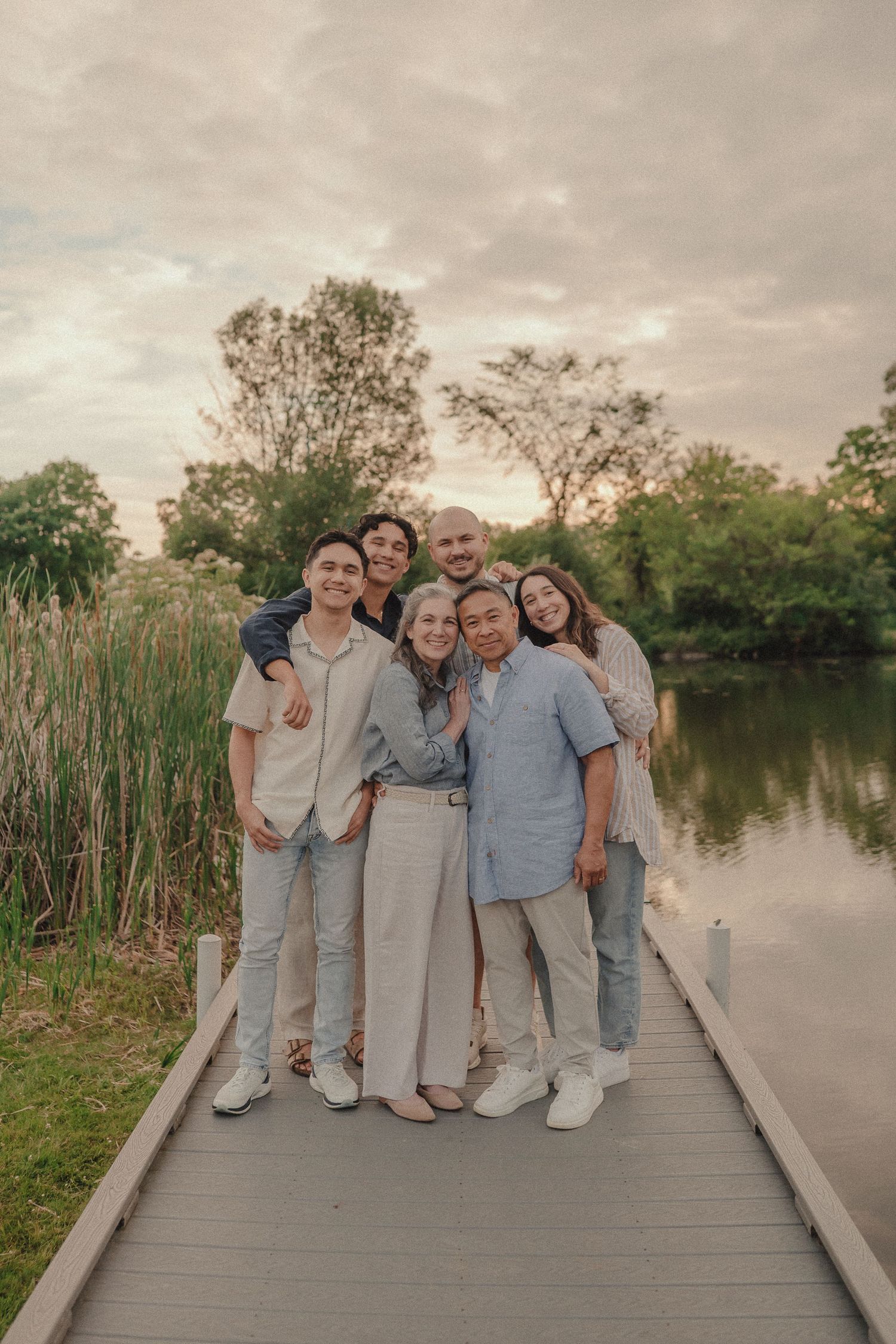 Happy multiracial extended family posing together on wooden dock by tranquil lake at sunset.