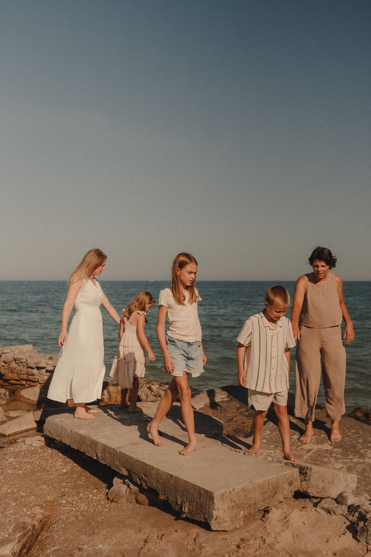 Family of five walking barefoot on rocky shore at sunset near calm blue sea.