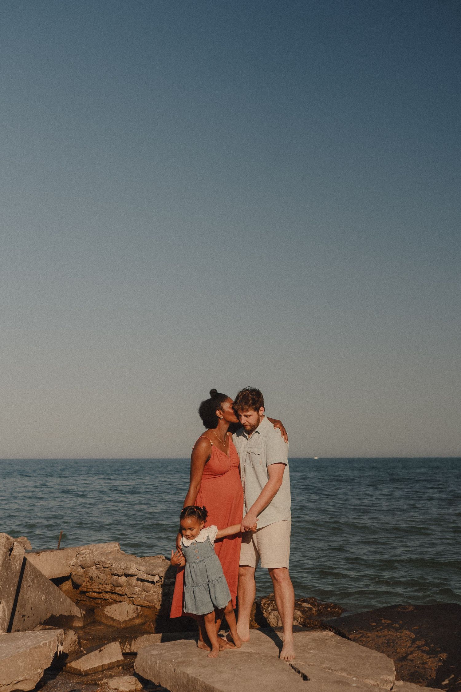 A family of three stands on rocky shoreline at sunset, with parents sharing a kiss while holding their young child's hands.