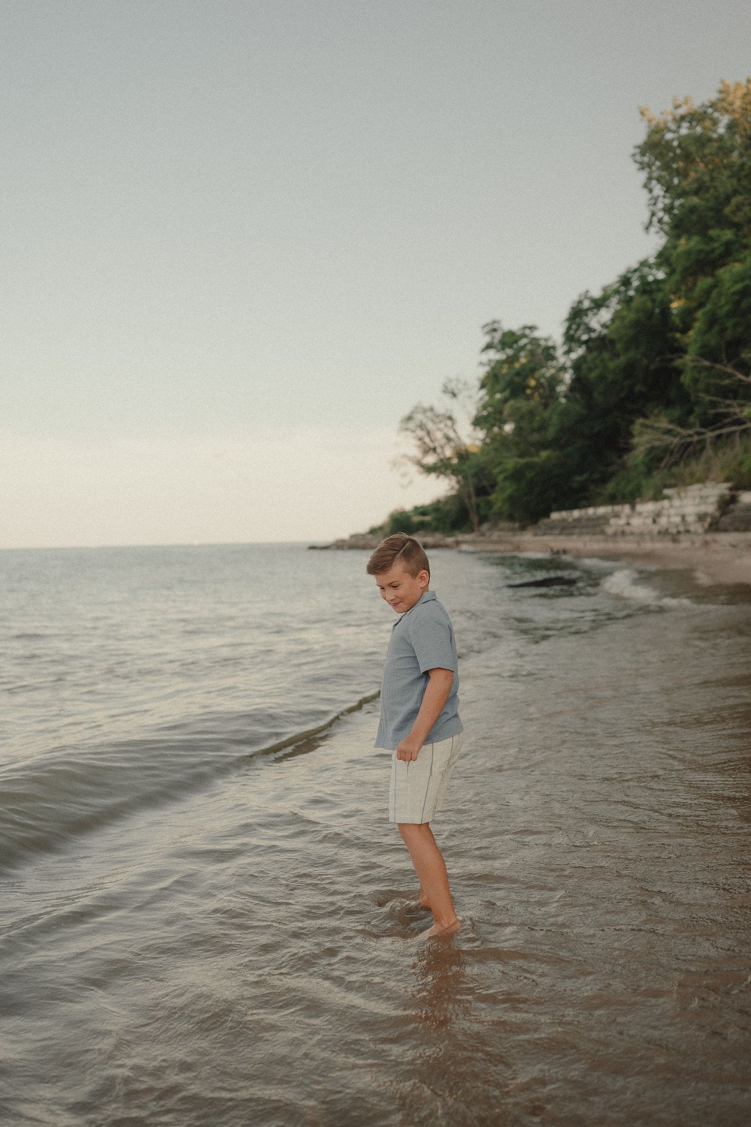 A young boy wades in shallow beach water, smiling back at the camera with trees and calm sea in the background.