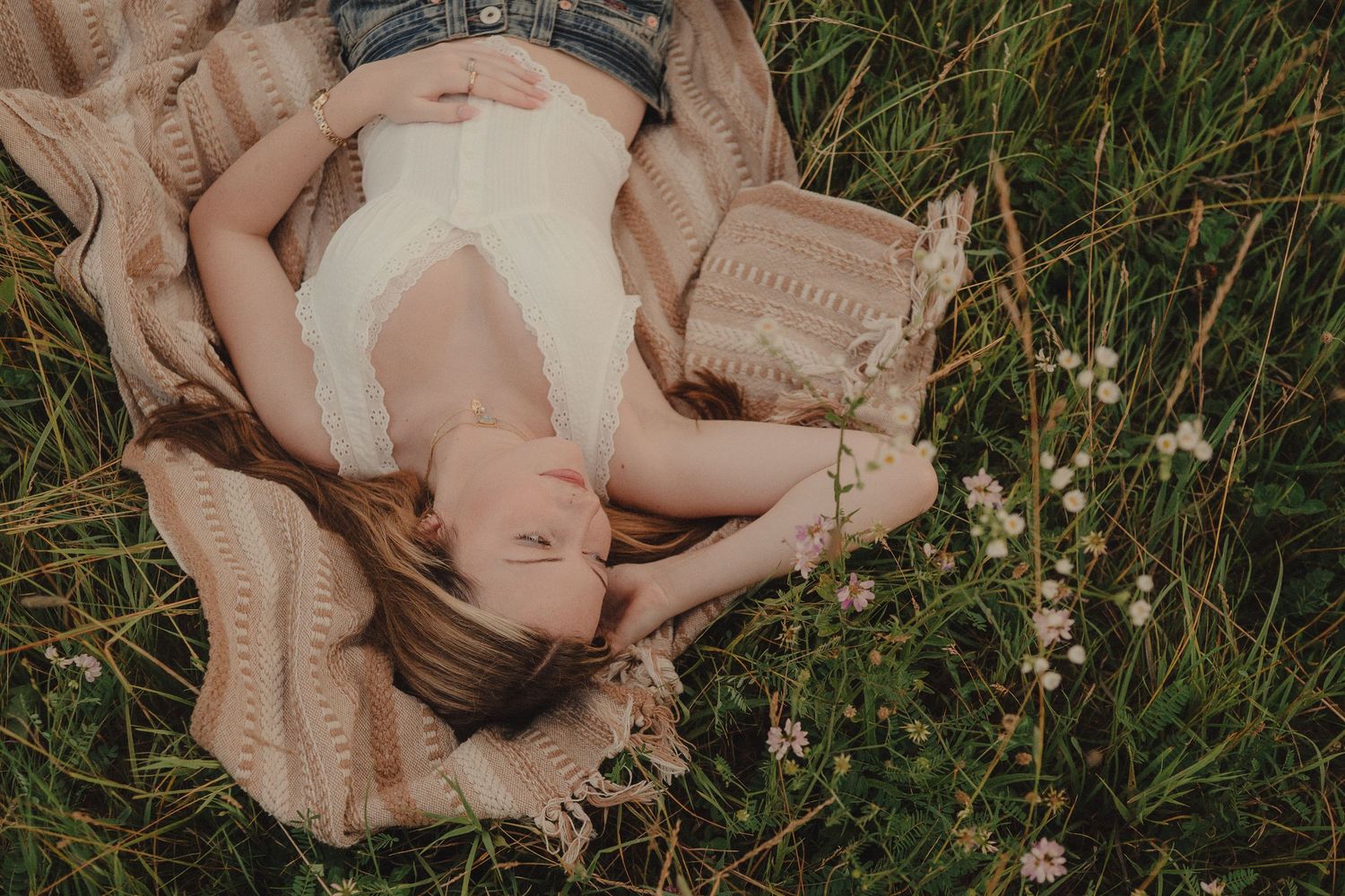 Woman lying on blanket in green meadow with wildflowers, relaxing outdoors on a sunny day.