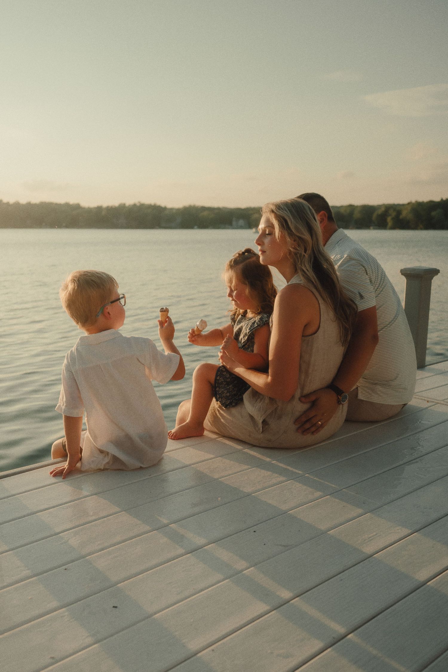 Family sits together on a wooden dock sharing popsicles at a peaceful lakeside sunset.