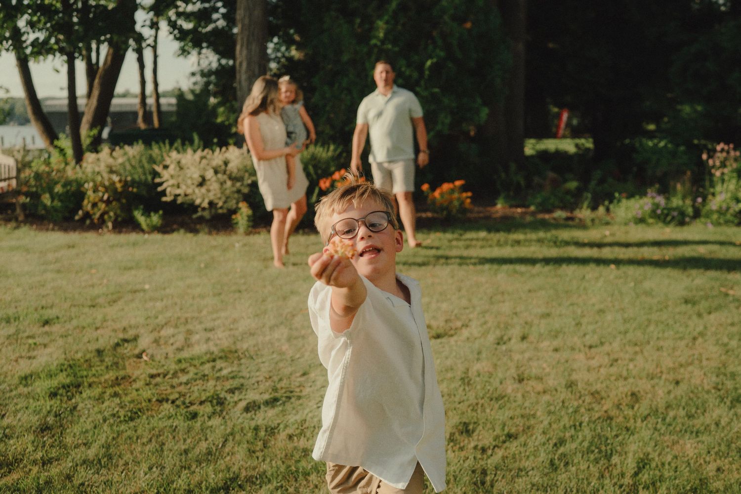 A young boy wearing glasses runs toward the camera in a sunny backyard while his parents play behind him.