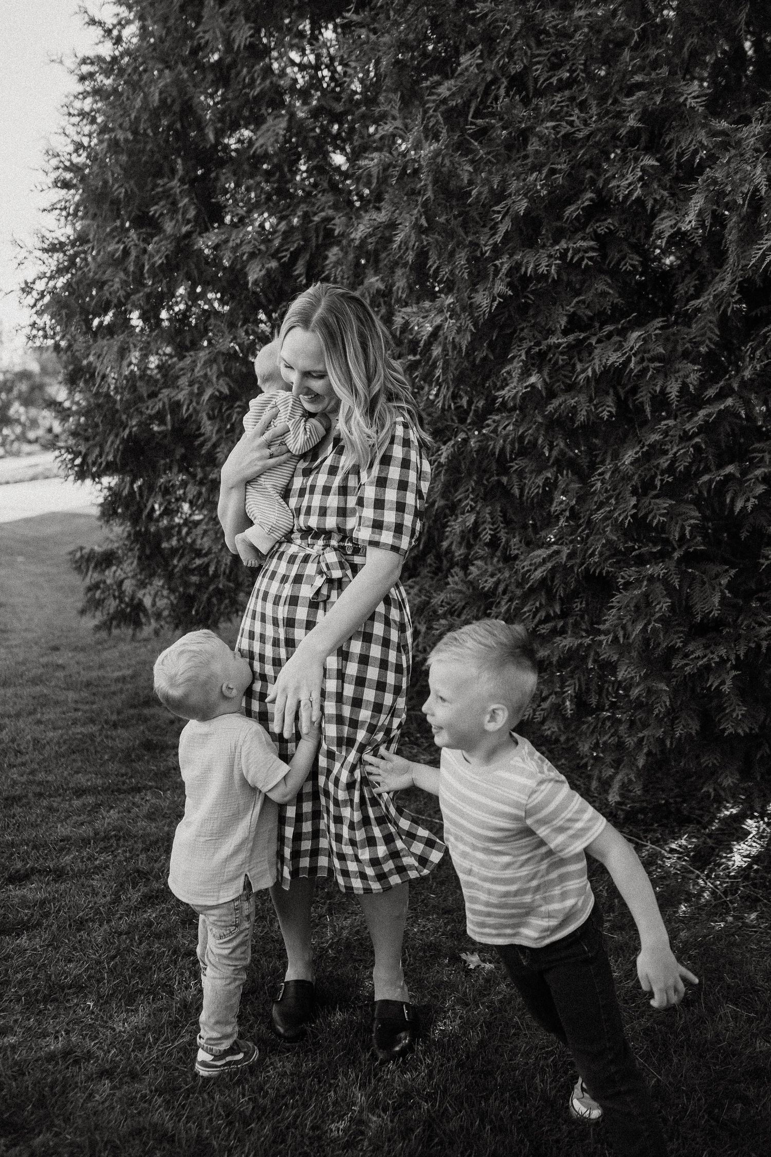 Pregnant woman in plaid dress laughing with three young children playing outdoors in black and white photo.