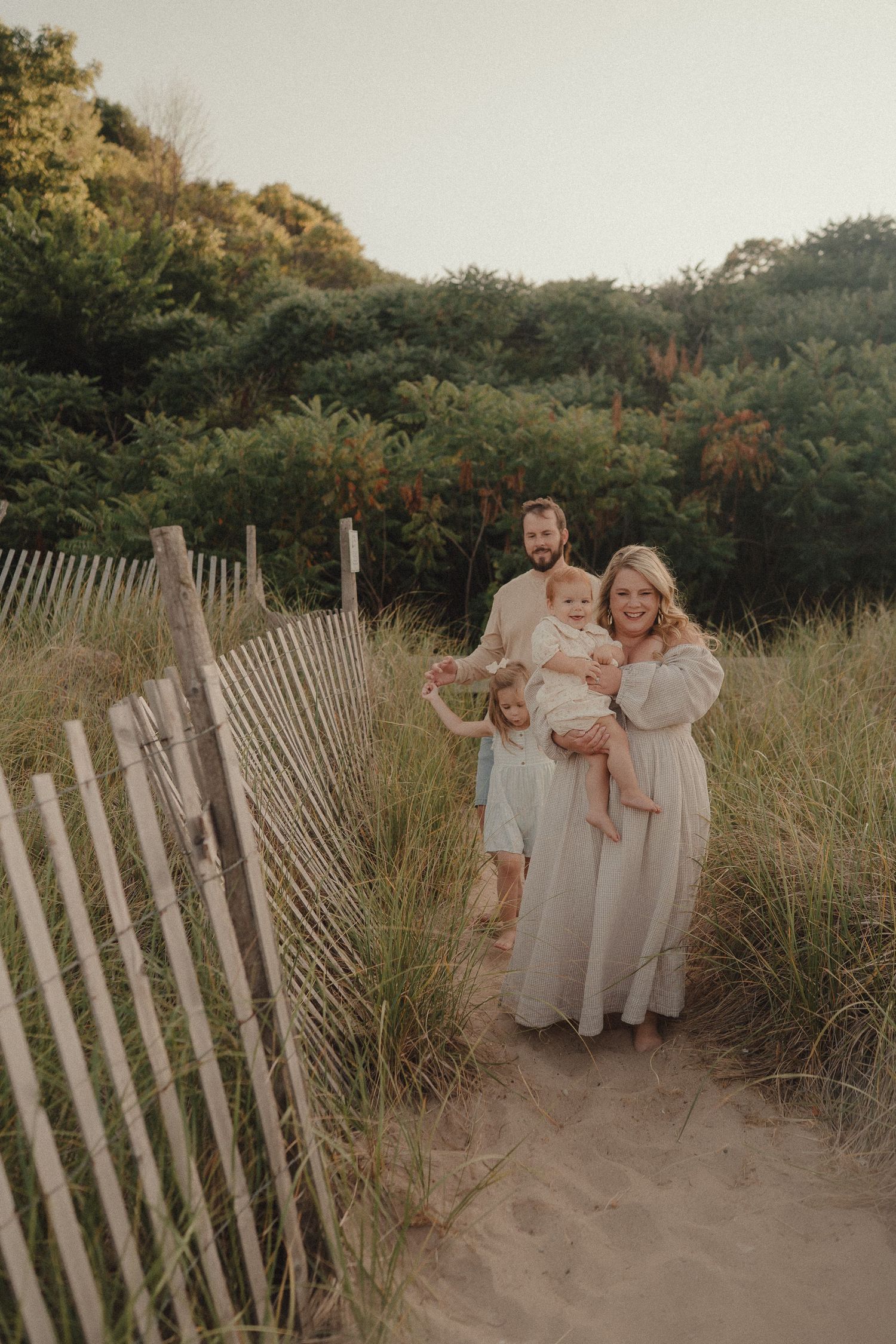 Family of four posing on sandy beach path lined with wooden fence and lush green dunes.