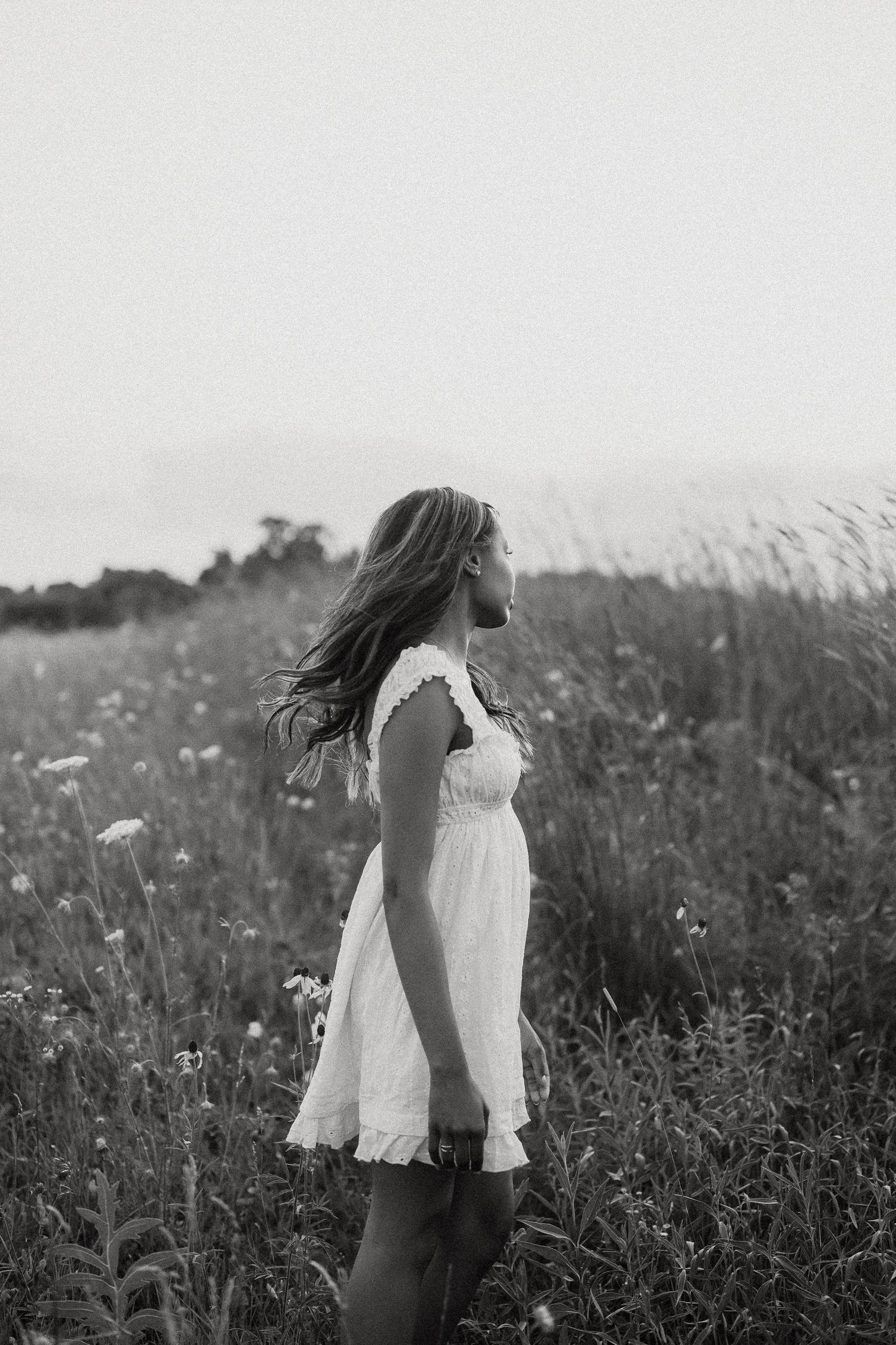 Black and white portrait of young woman in white dress standing in a wildflower meadow.