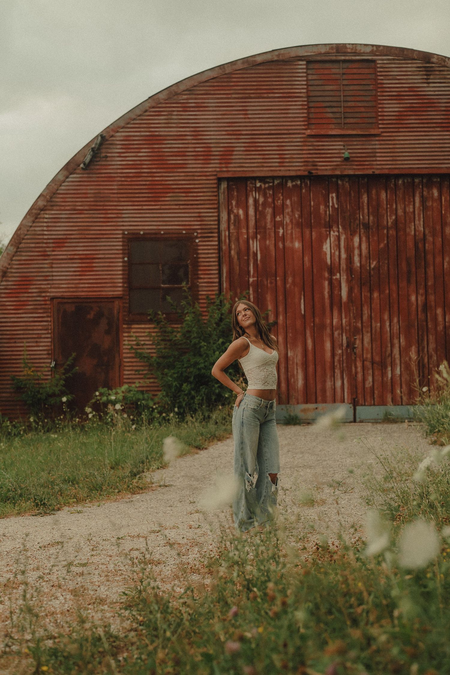 Young woman in white crop top and ripped jeans posing in front of a rustic red corrugated metal barn.