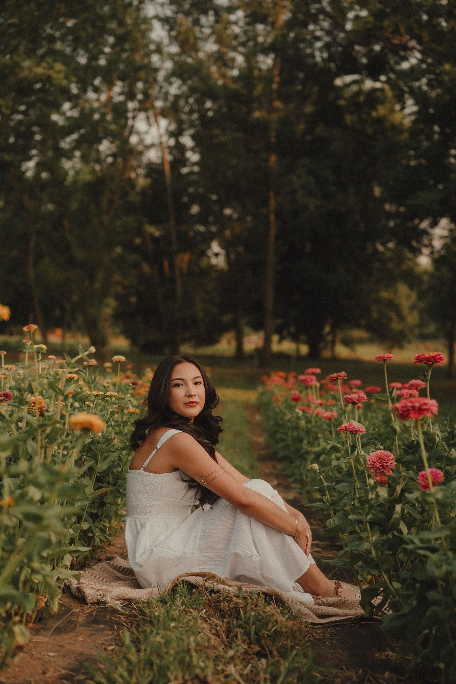 Young woman in white dress sitting among colorful dahlias in a garden at sunset.