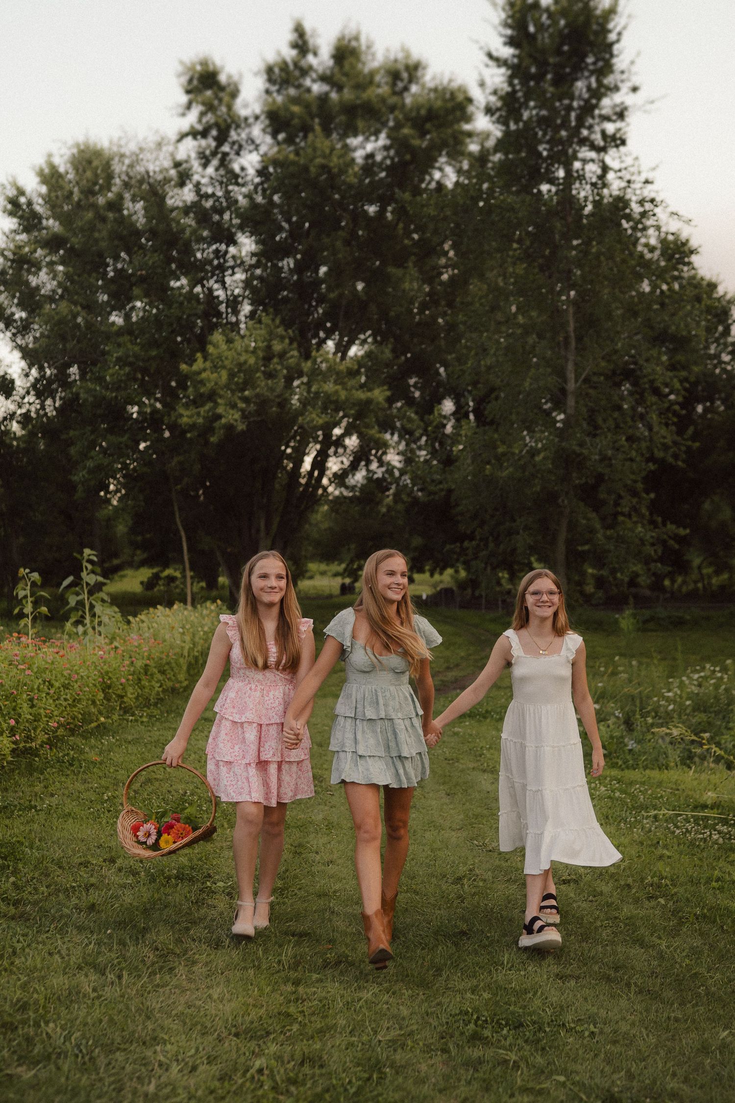 Three people in flowy summer dresses walk together through a sunlit grassy field on a warm afternoon.
