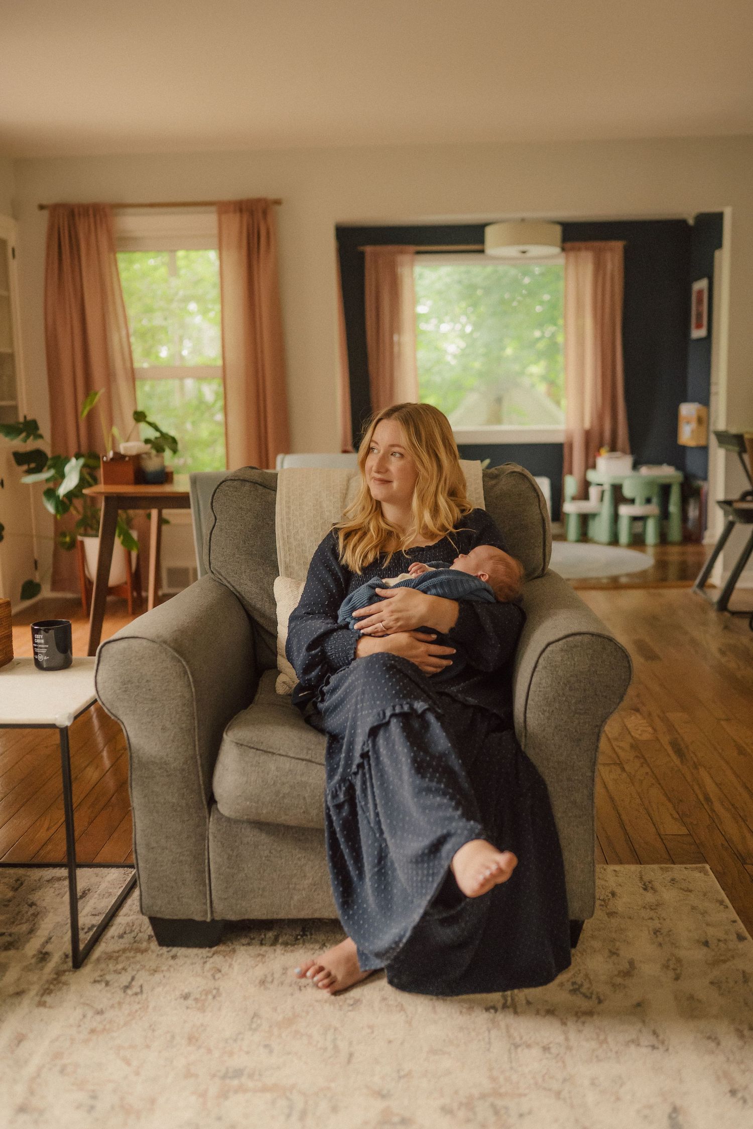 Warm family scene in sunlit living room as new parents bond with infant on comfortable grey armchair.