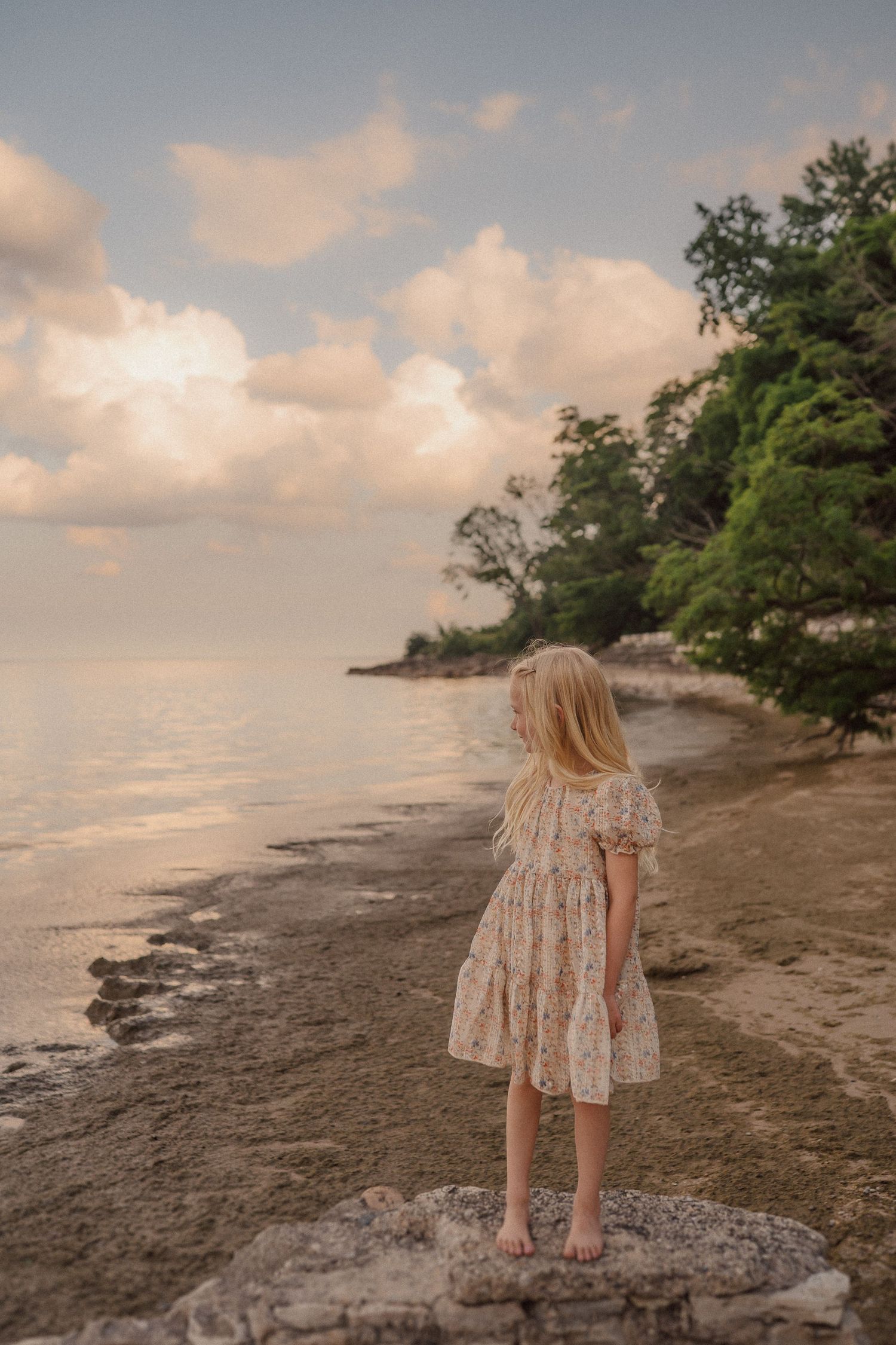 A young girl in a floral dress stands on a rocky beach during sunset while looking out at the tranquil water.