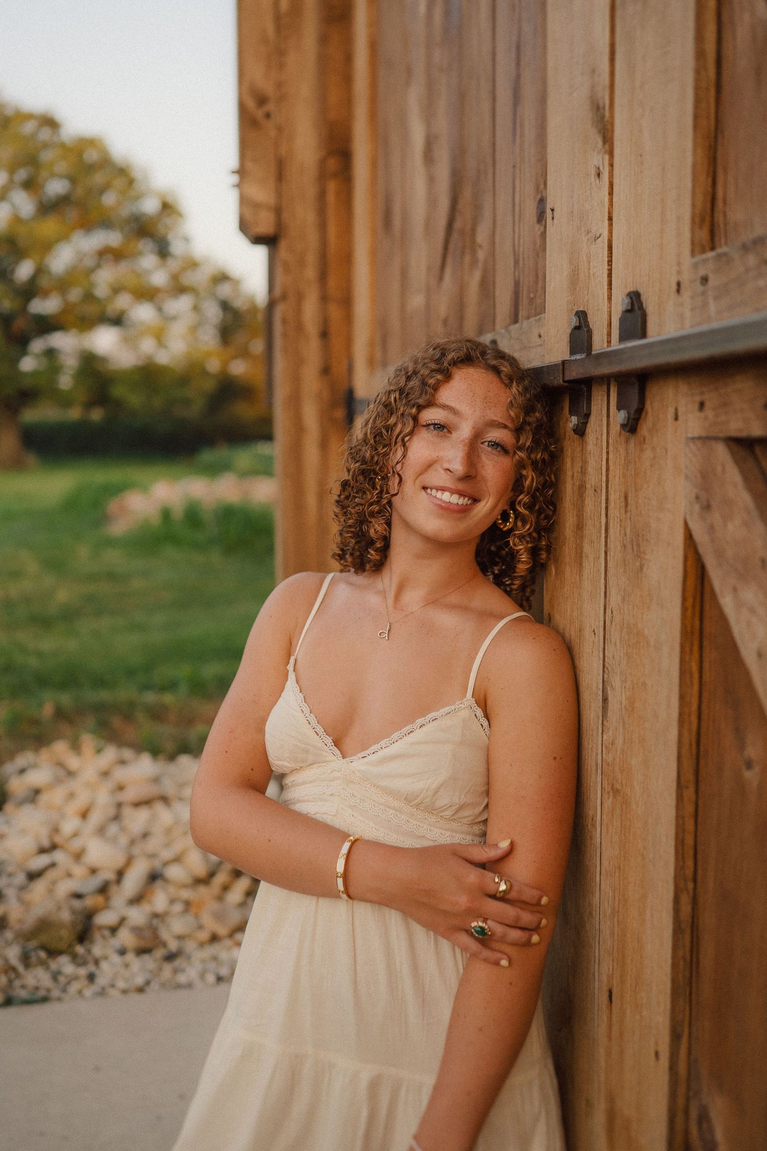 Someone wearing a cream-colored dress poses against a rustic wooden barn wall at sunset.