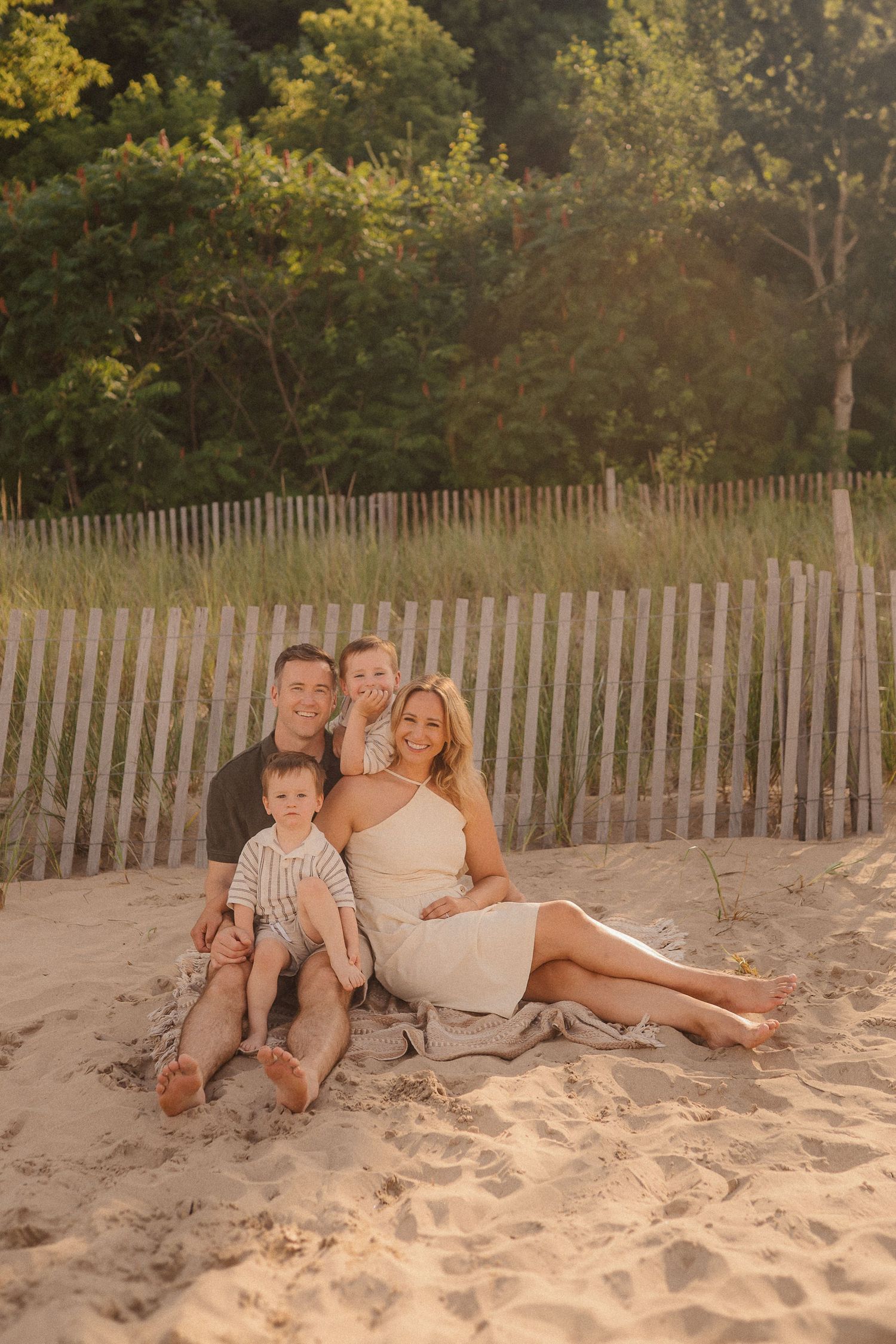A family sits together on a sandy beach at sunset with a wooden fence and trees in the background.